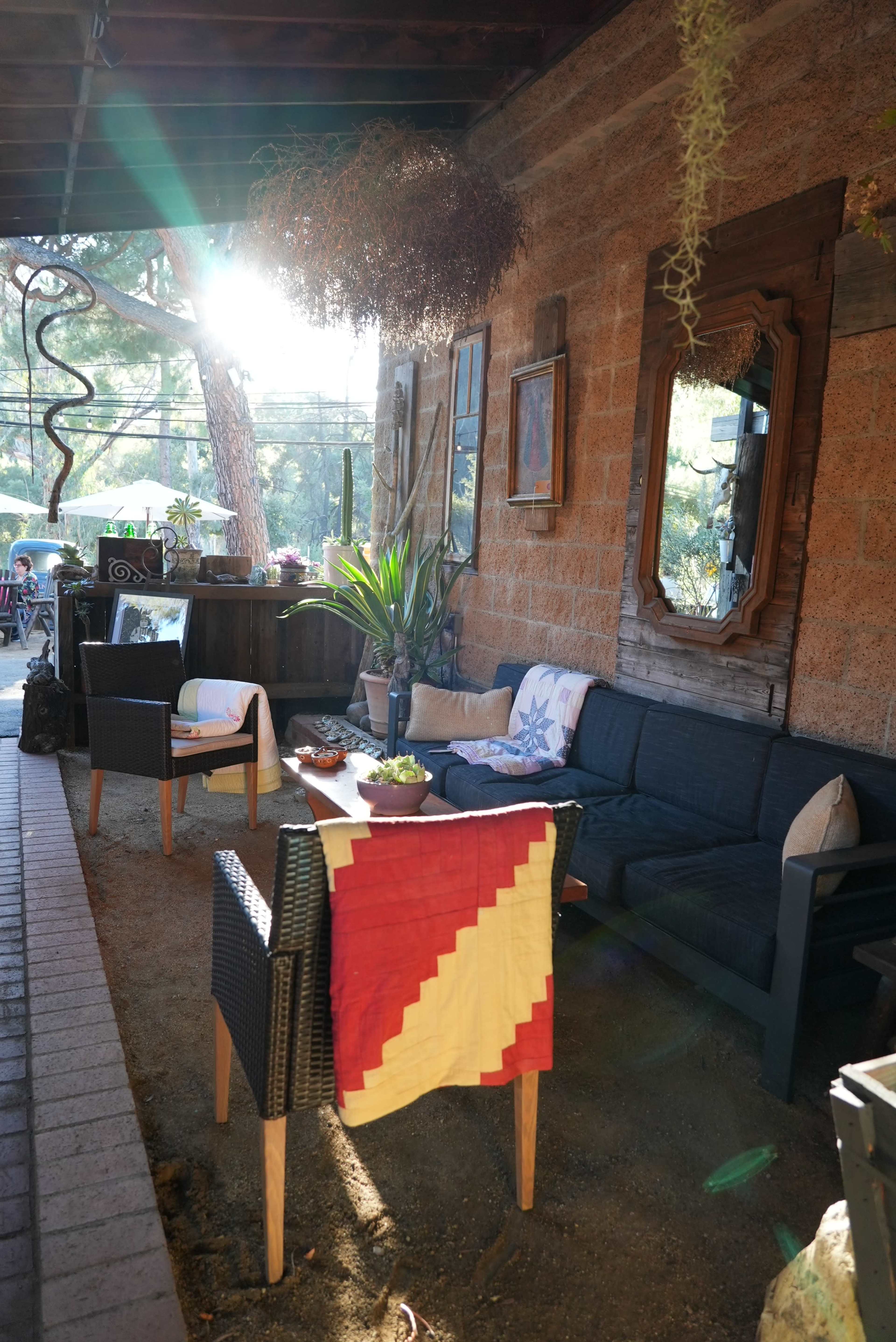 A cozy outdoor seating area features a black sofa, matching chairs, and a small table adorned with plants, set against a rustic wall illuminated by sunlight.