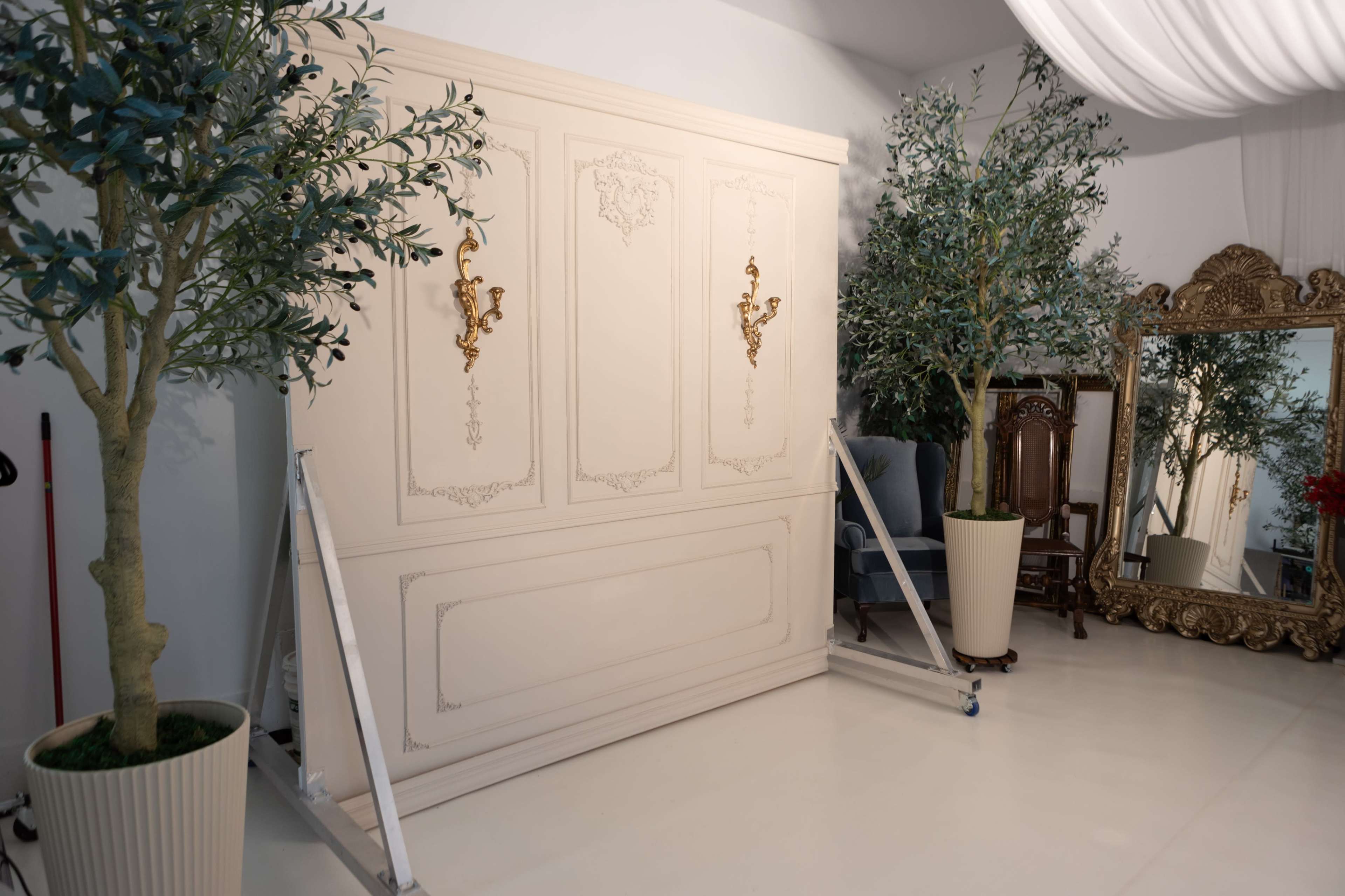 The image shows a well-decorated room with a large white cabinet featuring gold accents, flanked by two potted olive trees and a vintage-style mirror.