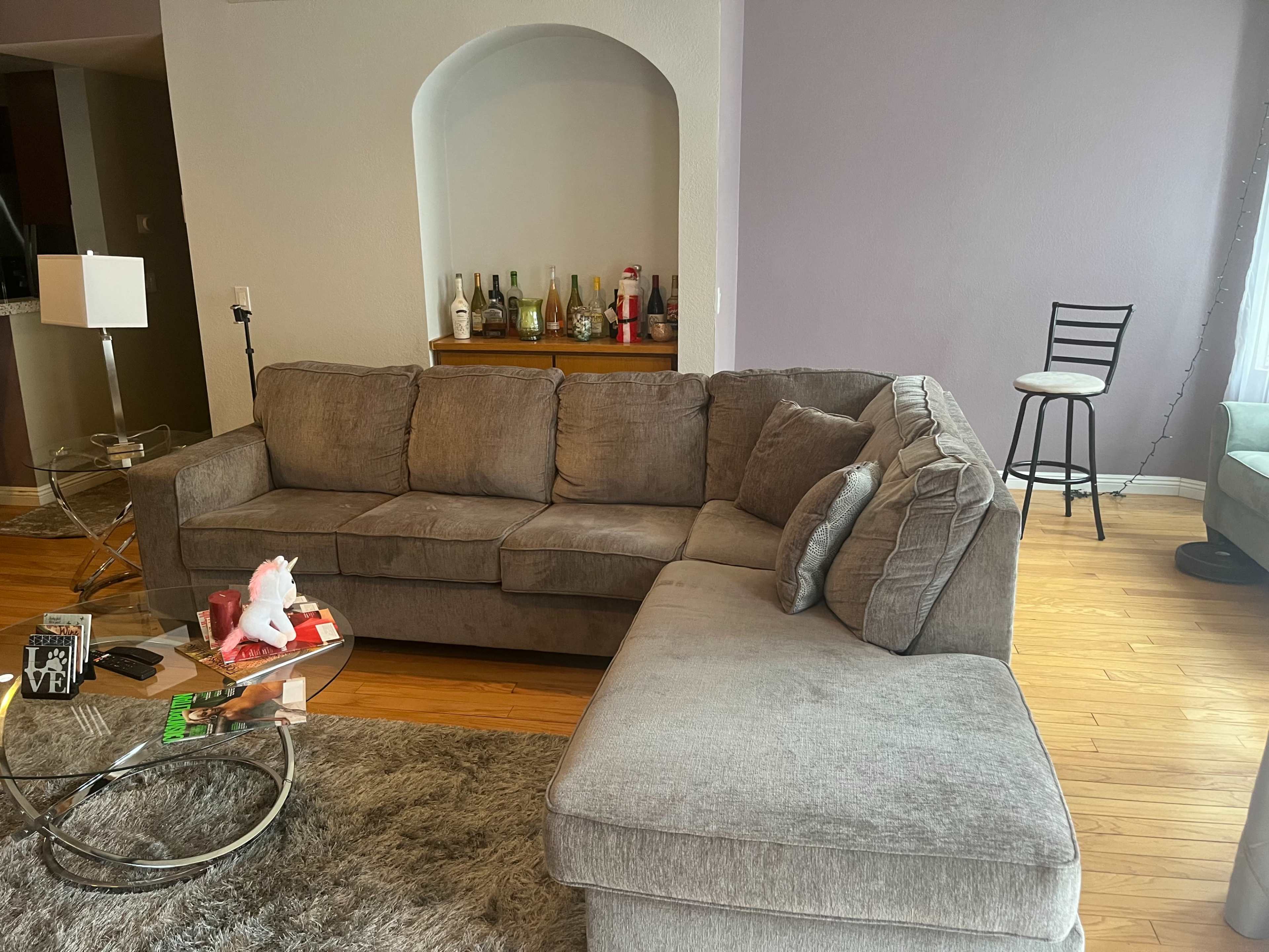 A gray sectional sofa is positioned in a living room with wooden flooring, a glass coffee table, and a wall shelf displaying several bottles.