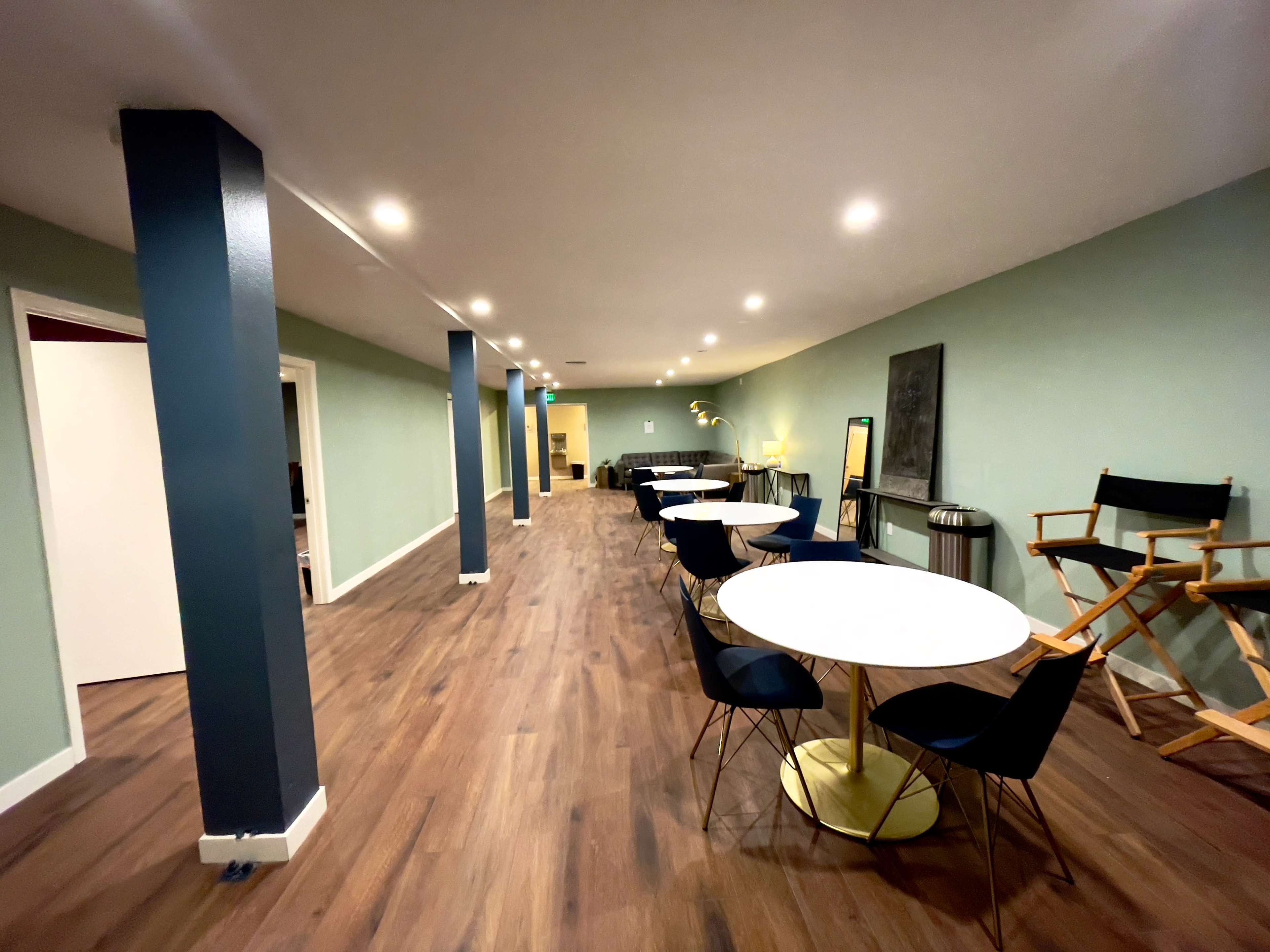 A long, empty room with wooden flooring, featuring several round tables and chairs, and light green walls with overhead lighting.