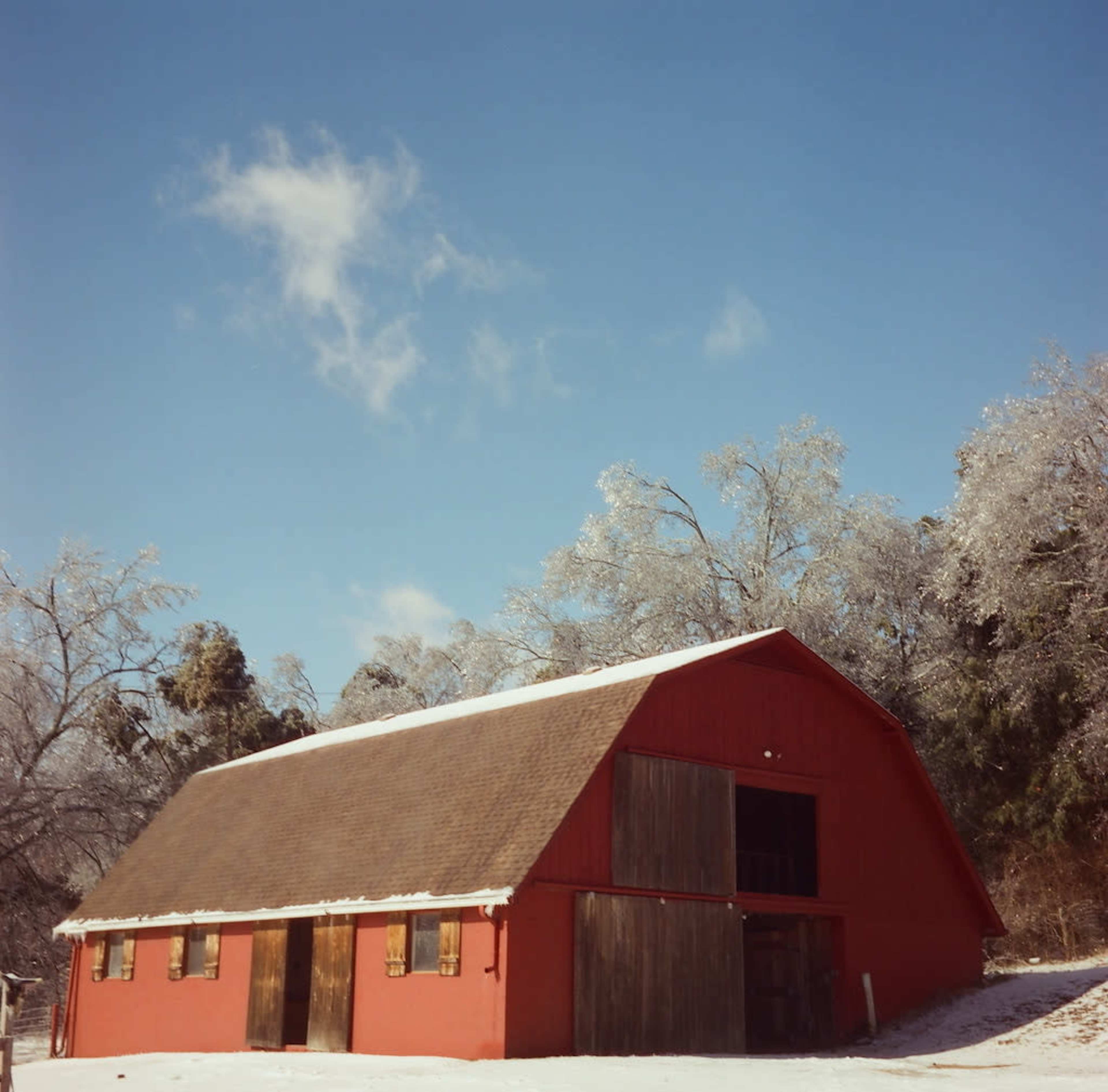 A red barn sits on a snow-covered landscape under a clear blue sky, surrounded by trees coated in ice.