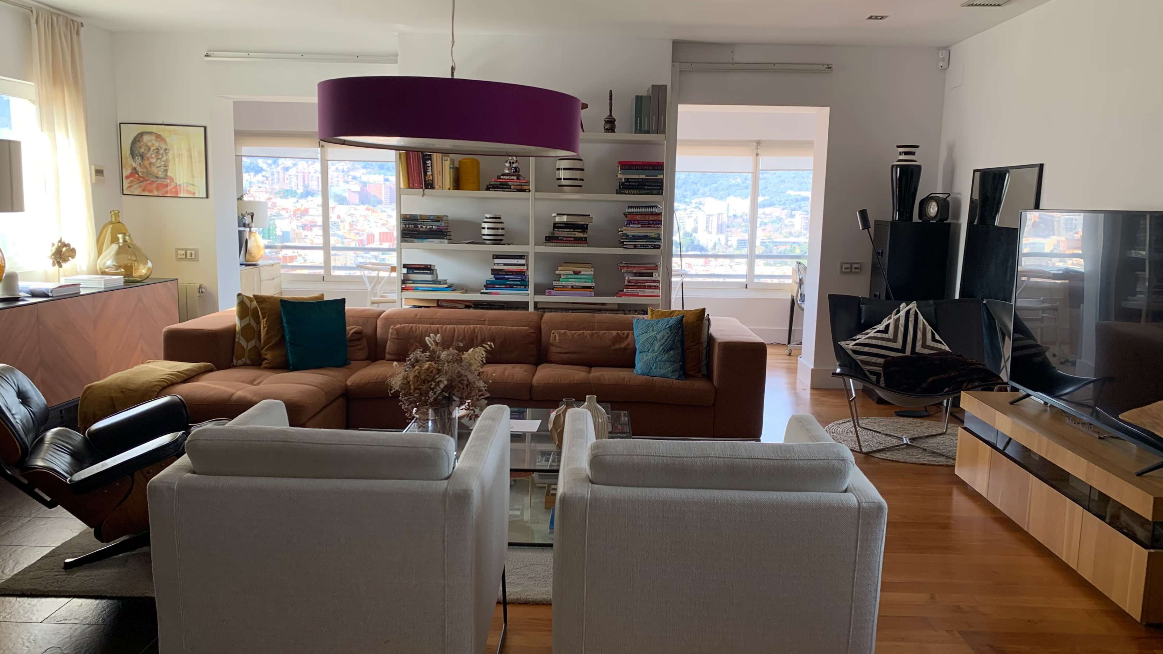 A modern living room features a brown sofa, white armchairs, a glass coffee table, and a bookshelf filled with books, all arranged under a purple pendant light.