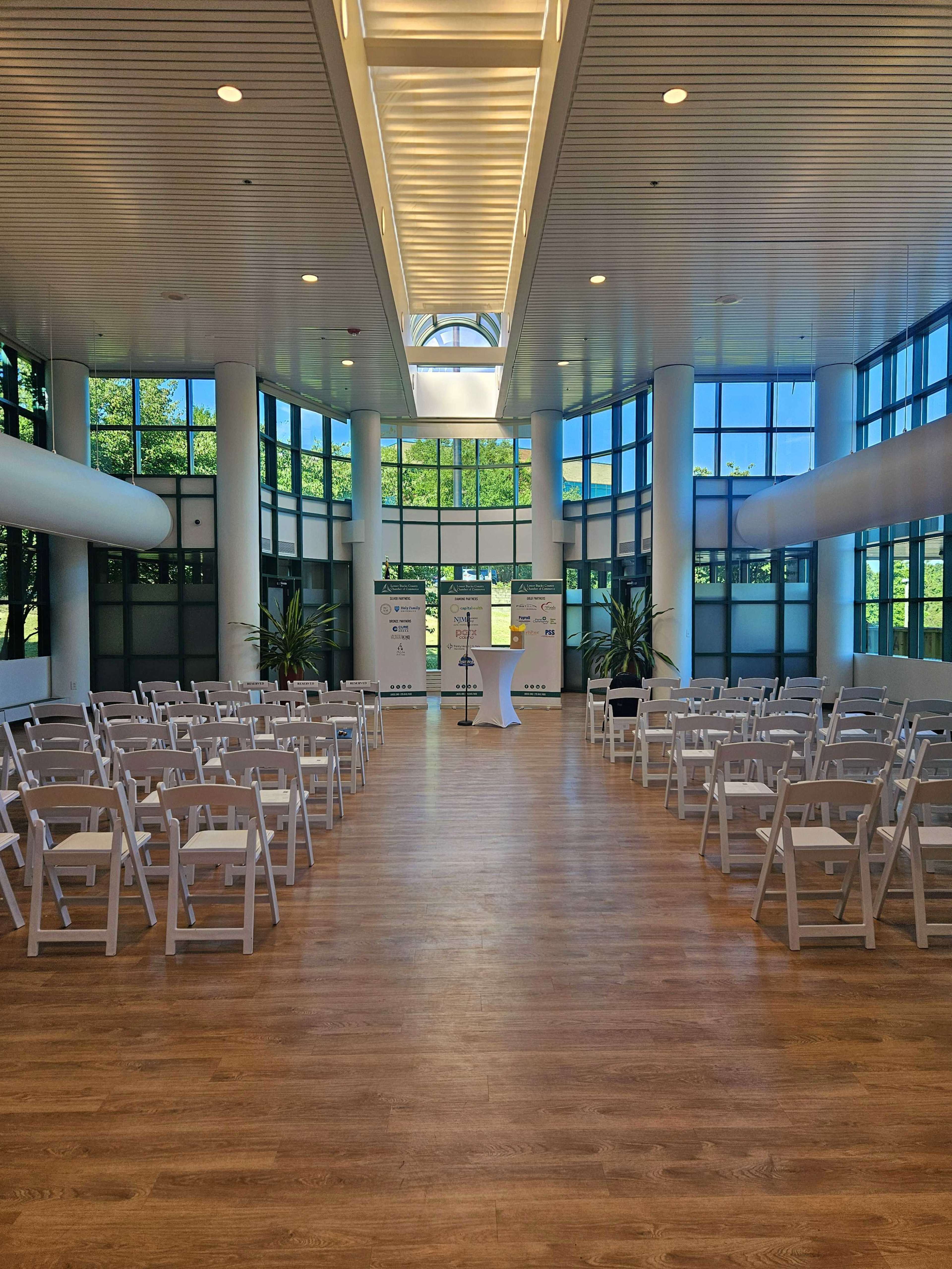 A large, bright indoor space with rows of white folding chairs facing a podium, surrounded by large windows and greenery.