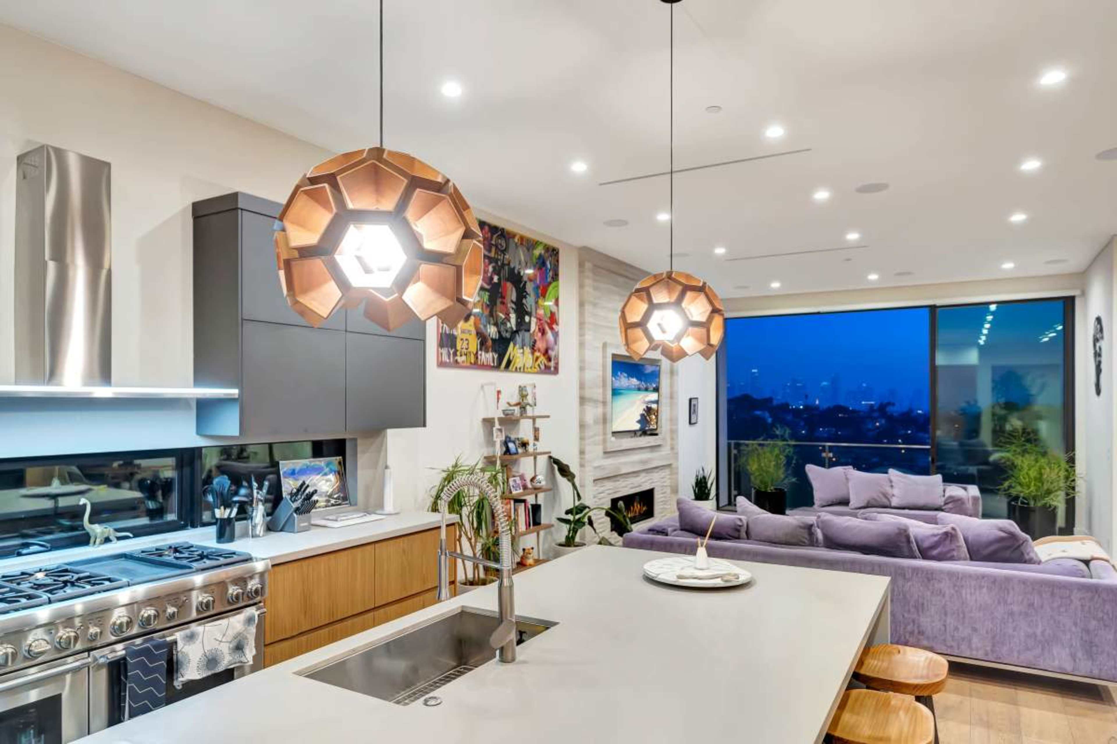 A modern kitchen features sleek cabinetry, a stainless steel appliance set, and pendant lighting, while a cozy living area with a purple sofa overlooks a city skyline through large windows.