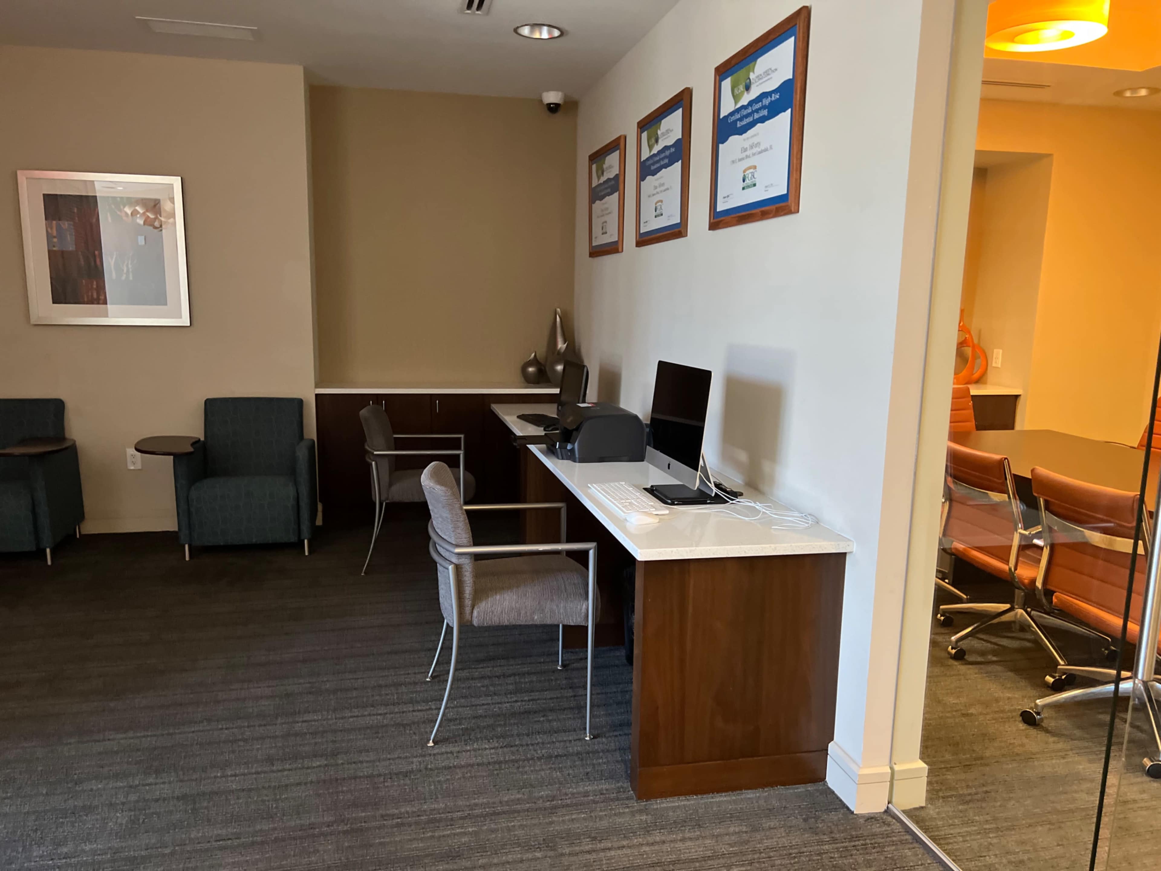 The image shows a small office area with a computer workstation, chairs, and framed certificates on the wall.
