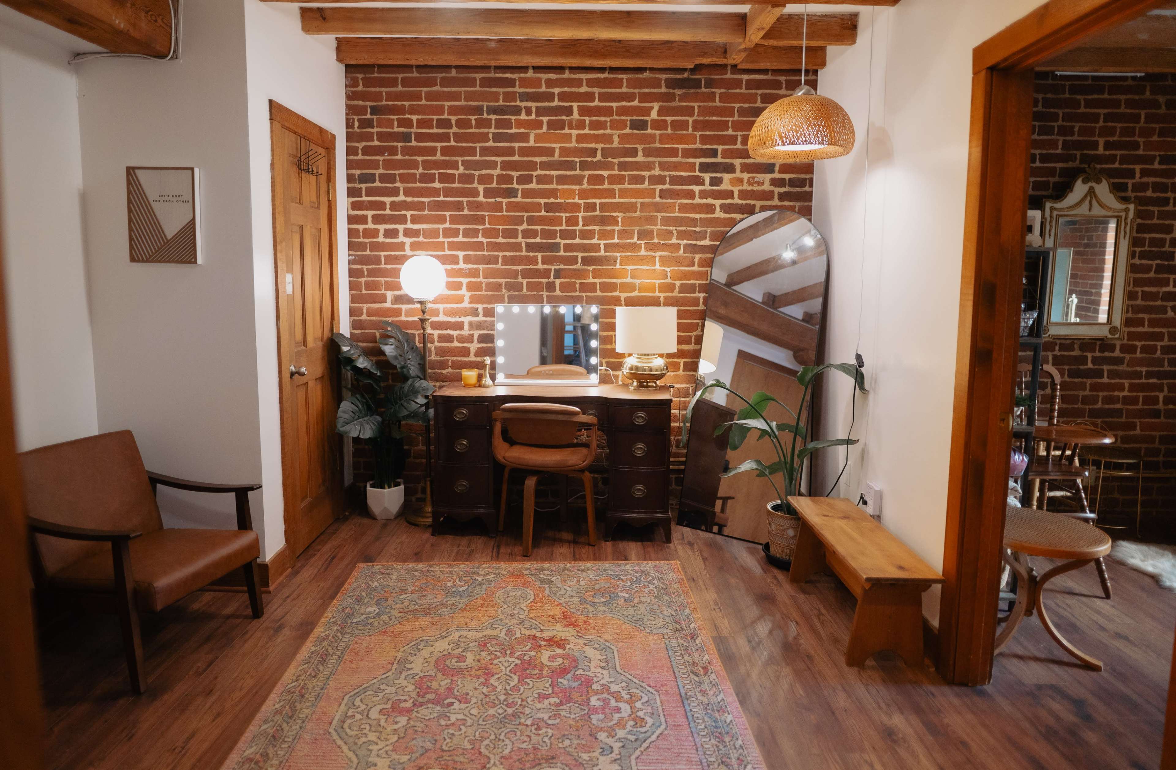 The scene features a cozy room with a brick wall, a wooden desk with a mirror, a lamp, and decorative elements like a plant and a vintage rug.