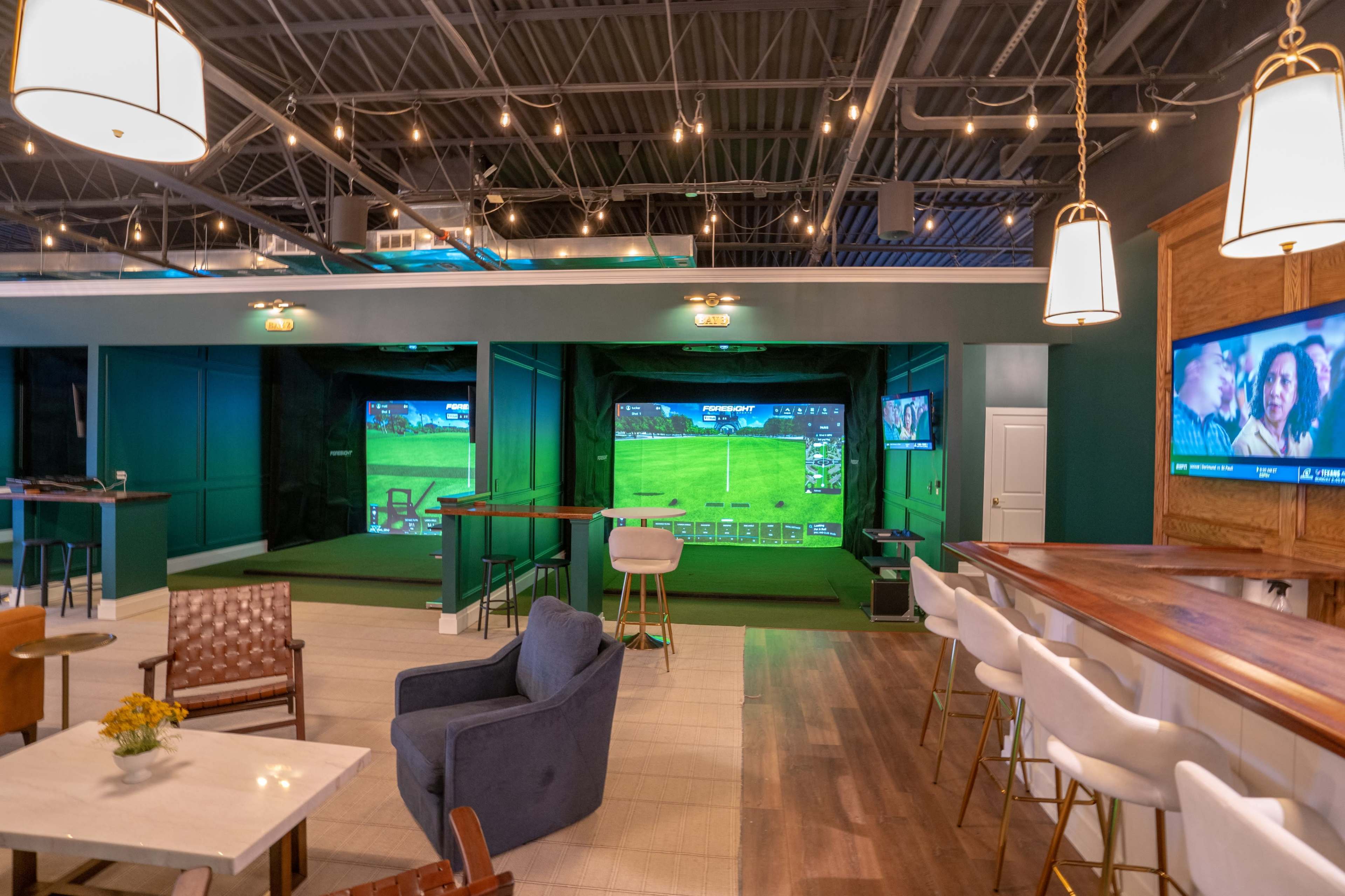 The image shows a modern indoor golfing area with two large screens displaying a golf course, surrounded by seating and tables in a well-lit space.