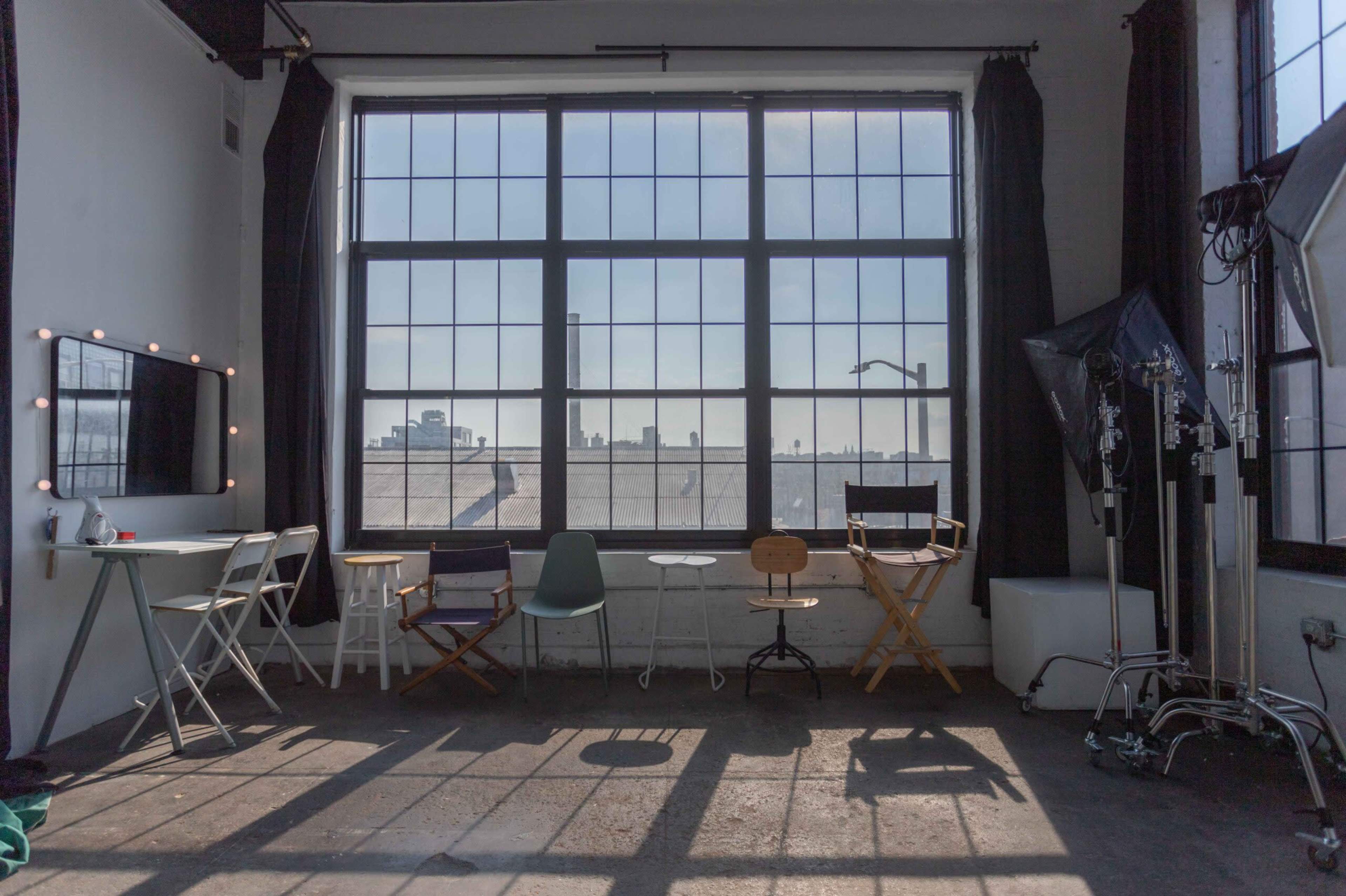 The image shows a spacious studio with large windows, several chairs, a mirrored makeup station, and photography equipment.