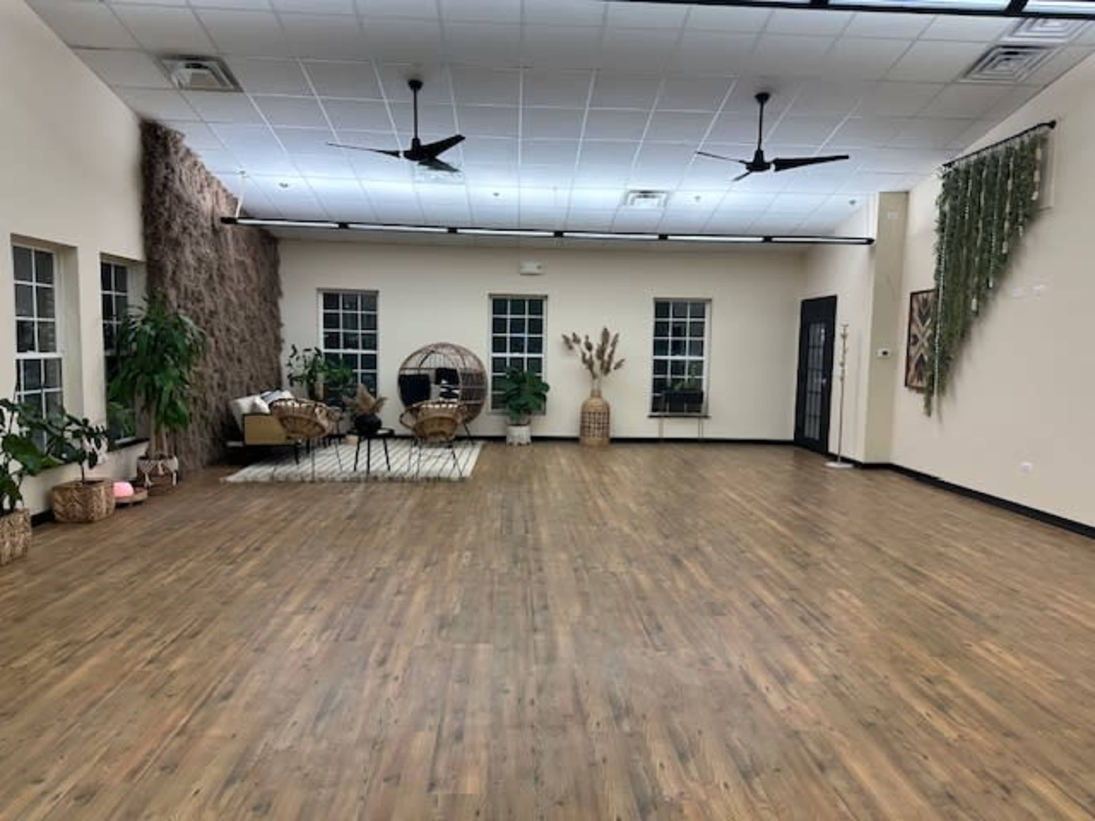 A spacious, empty room with wooden flooring, large windows, and various decorative plants, including a hanging wall feature and a seating area with wicker chairs.