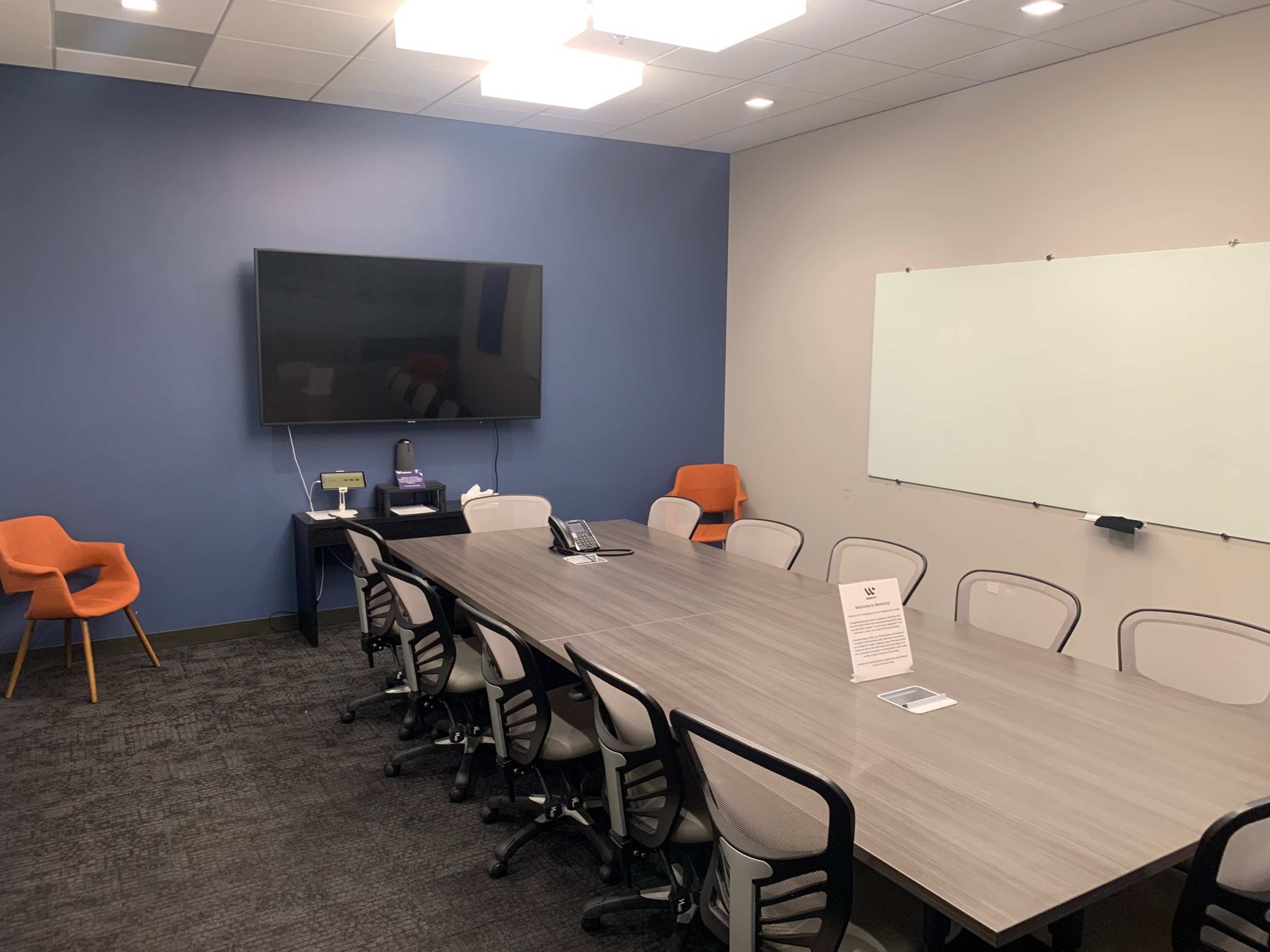 A conference room features a long table surrounded by chairs, a large wall-mounted TV, an orange armchair in the corner, and a whiteboard.