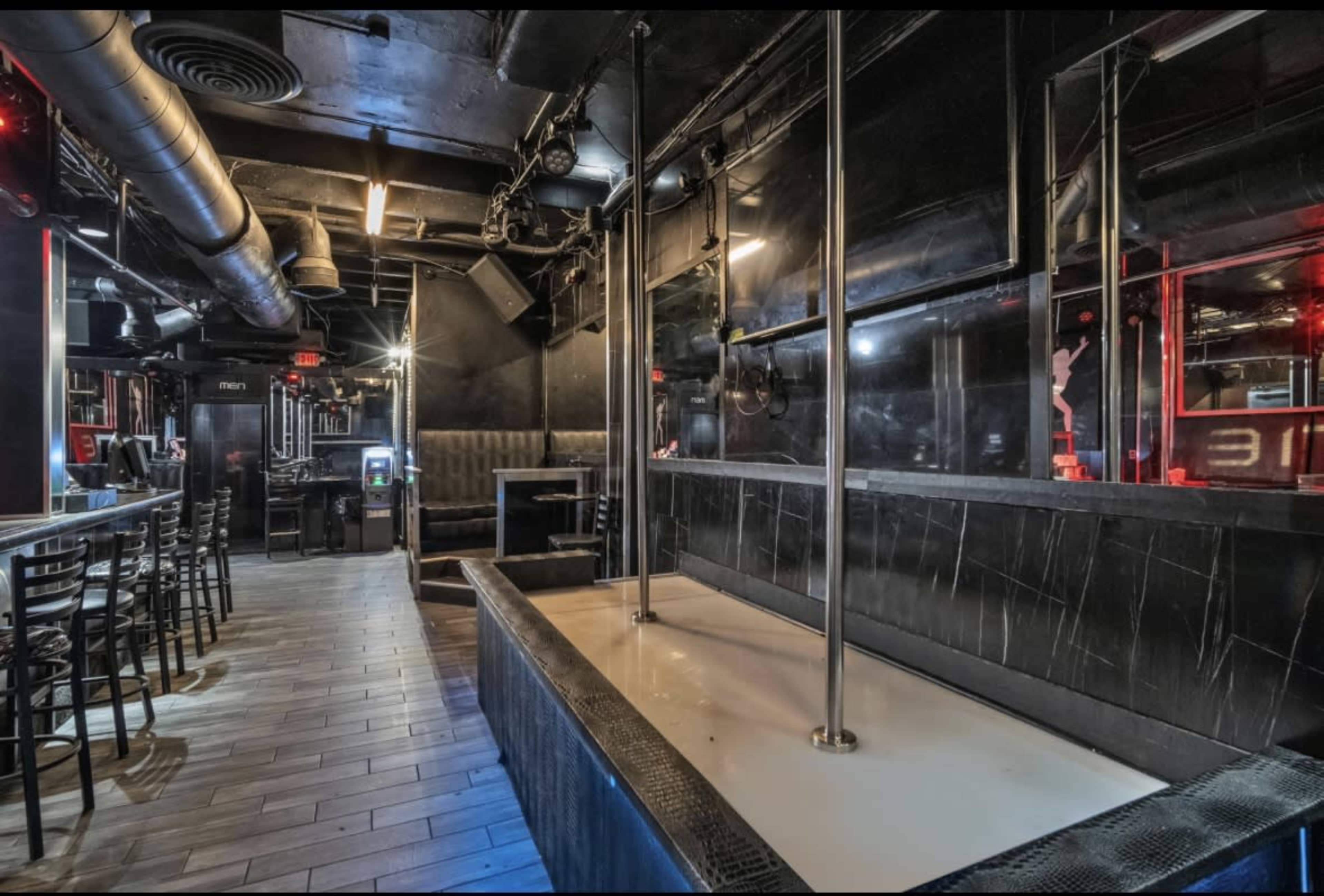 The image shows the interior of a dimly lit nightclub featuring a bar area, seating, a dance floor with a pole, and mirrored walls.