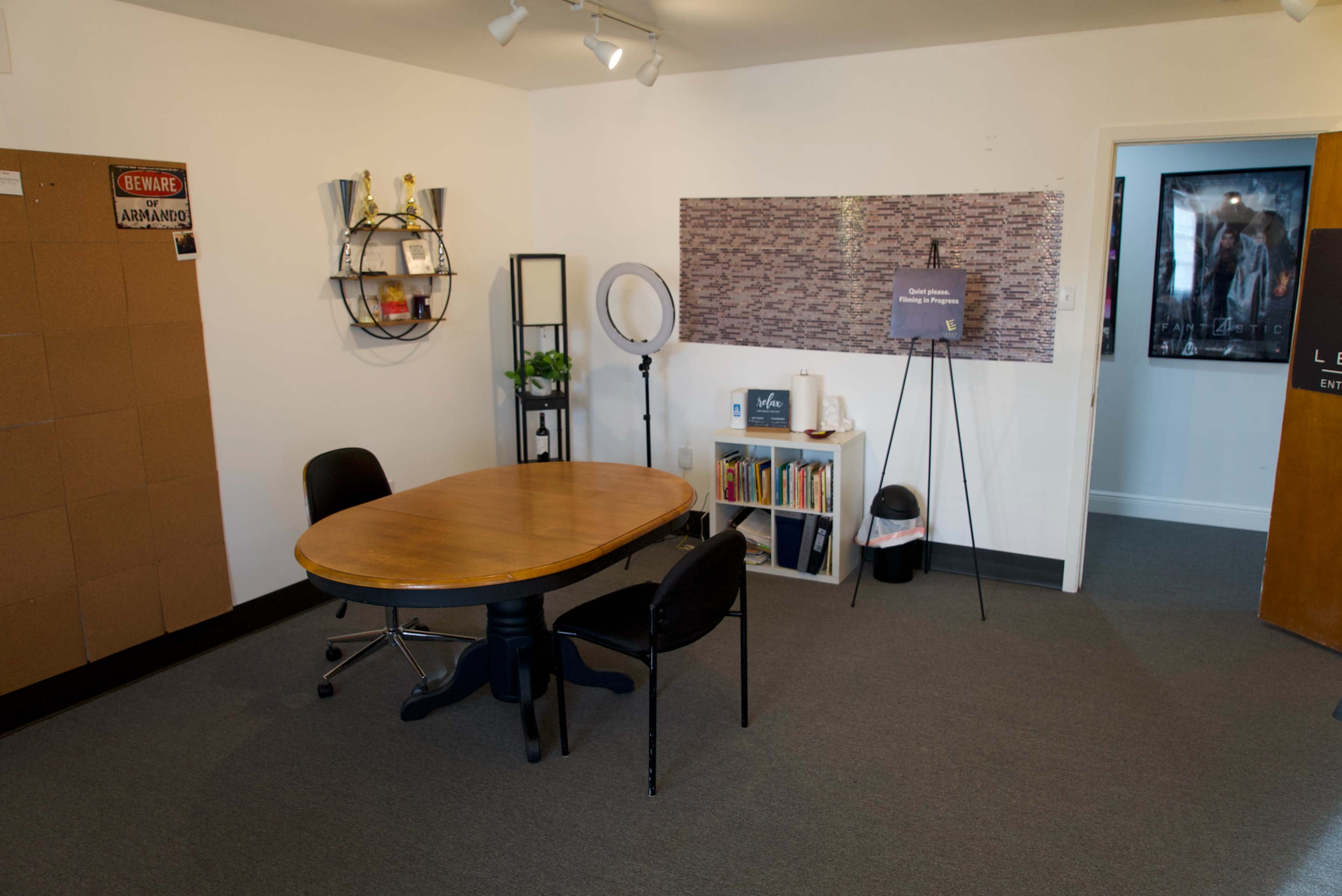 A minimalistic office space featuring a wooden round table with two black chairs, a bookshelf, a plant, and a board with pinned items on the wall.