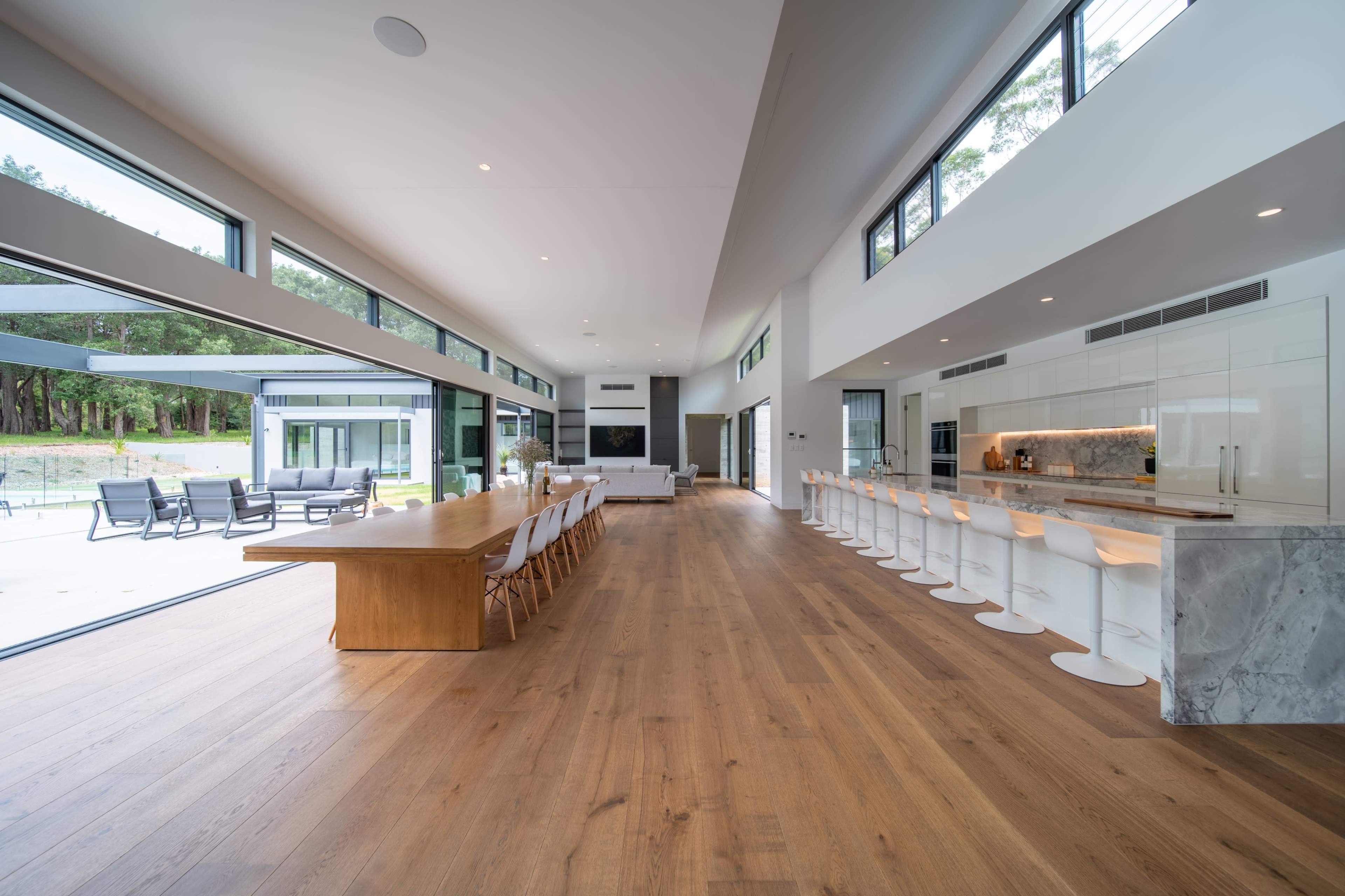 A modern, open-concept living space featuring a long wooden dining table, white bar stools, and large windows that overlook a patio and wooded area.