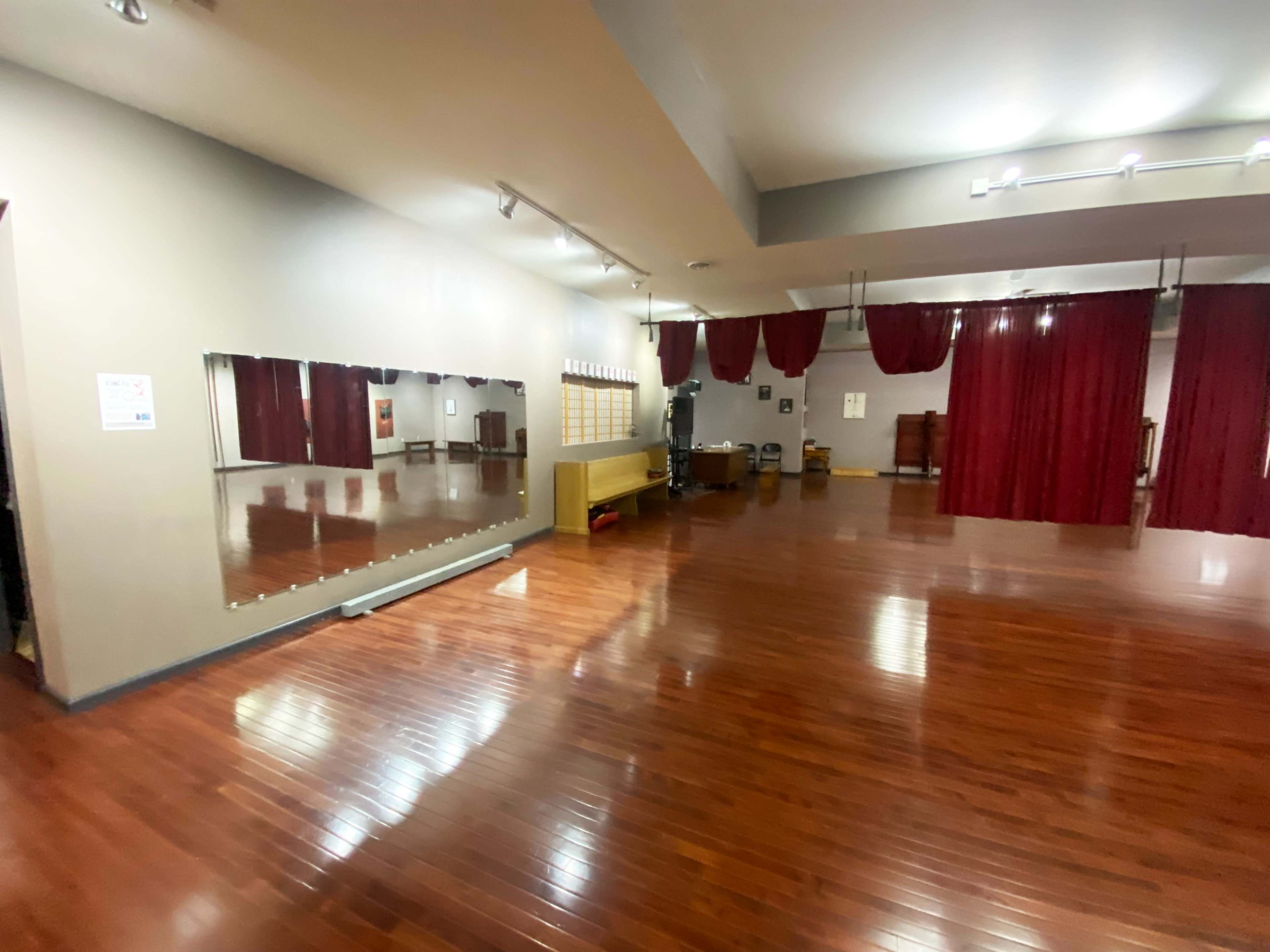 The image shows a spacious dance studio with polished wooden floors, a large mirror along one wall, and red curtains in the background.