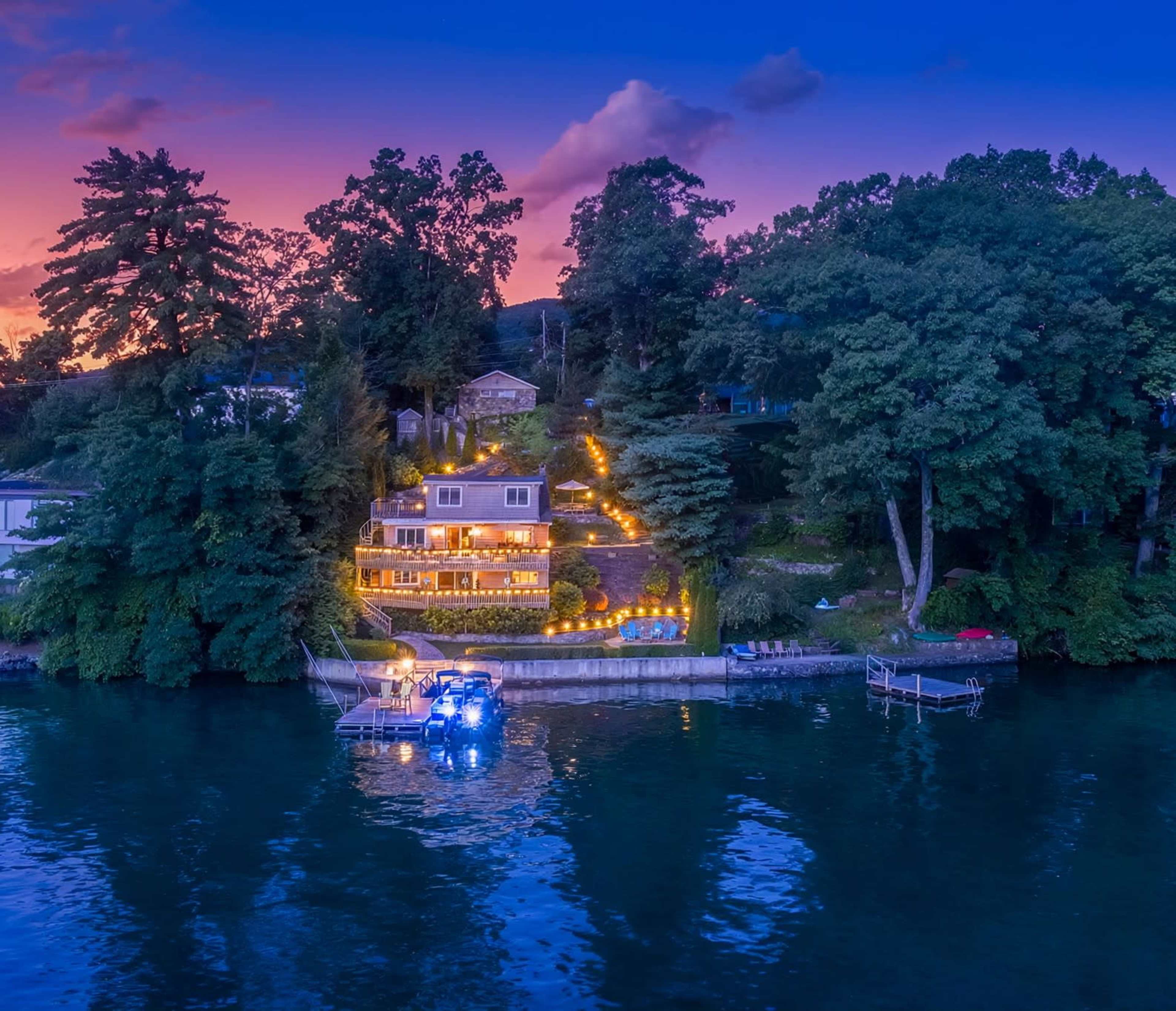 A waterfront house with outdoor lighting is situated on a grassy slope beside a calm lake, surrounded by trees, as twilight sets in.