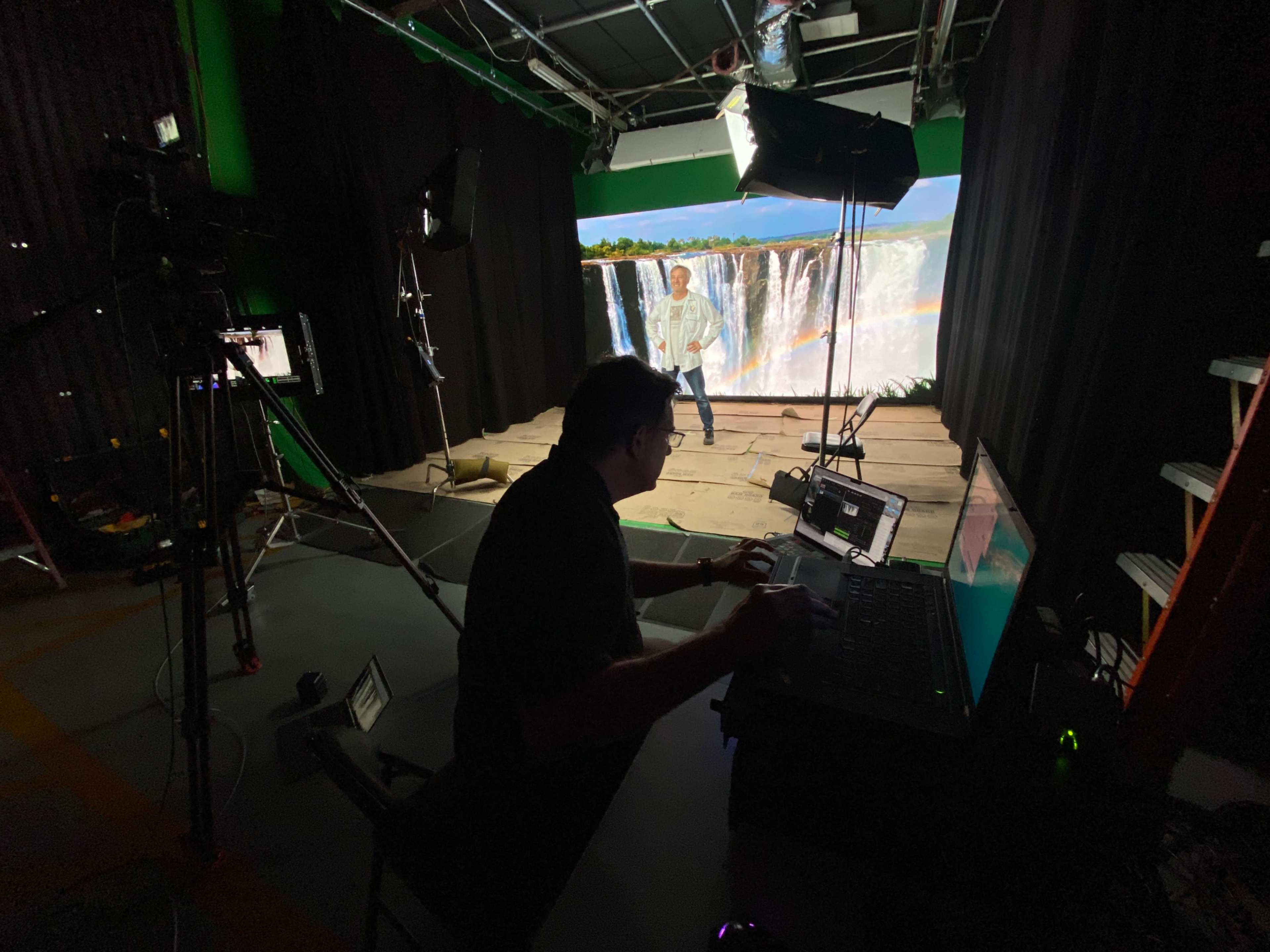 A person operates a computer at a workspace in a studio while a presenter stands in front of a large green-screen backdrop depicting a waterfall.