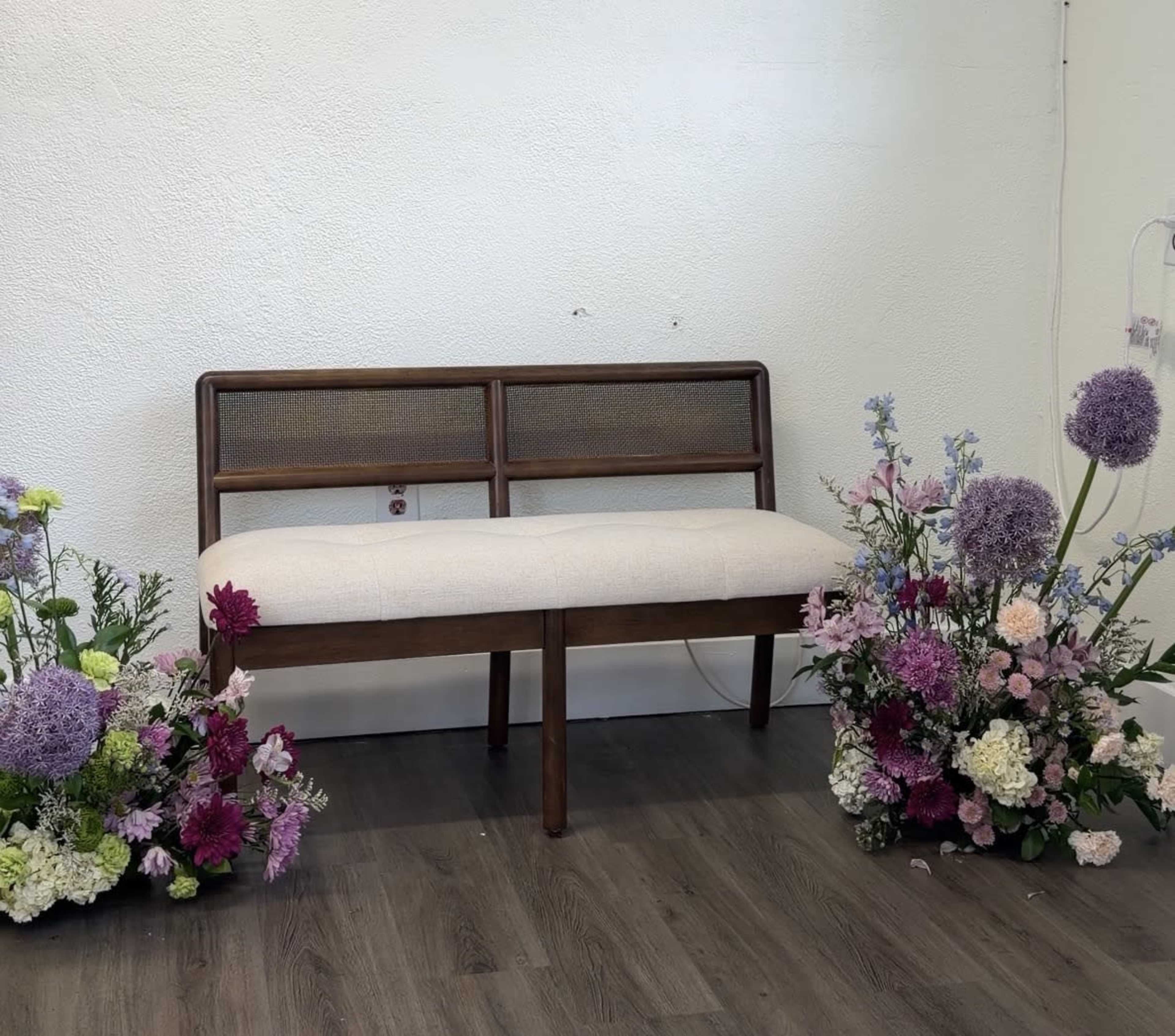 A wooden bench is positioned against a plain wall, surrounded by an arrangement of colorful flowers on either side.