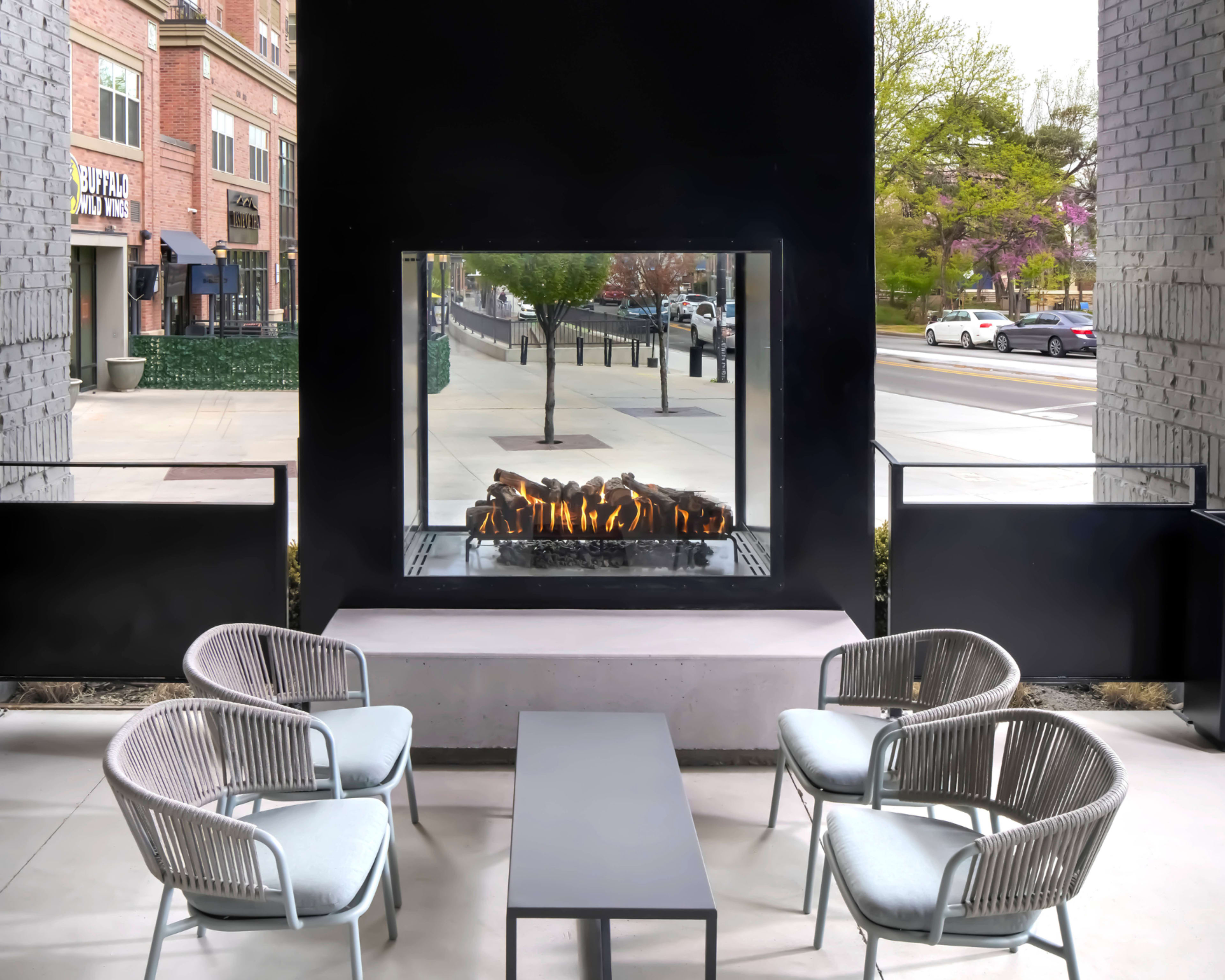 The image shows a modern outdoor seating area with four stylish chairs and a central table, featuring a large square fireplace that offers a view of flames through a glass panel.