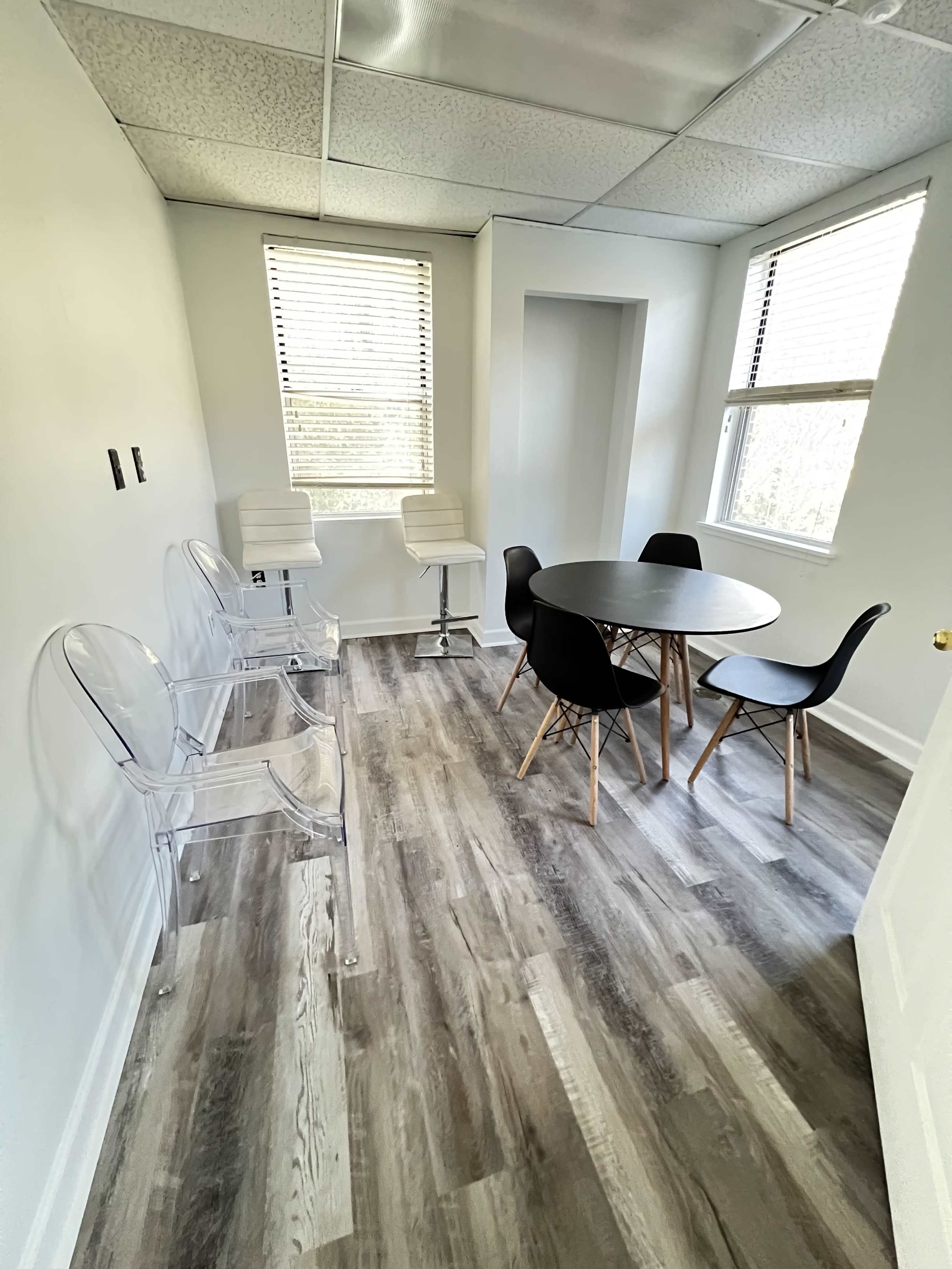 A bright, minimalist meeting room with a round black table surrounded by black and transparent chairs and large windows.