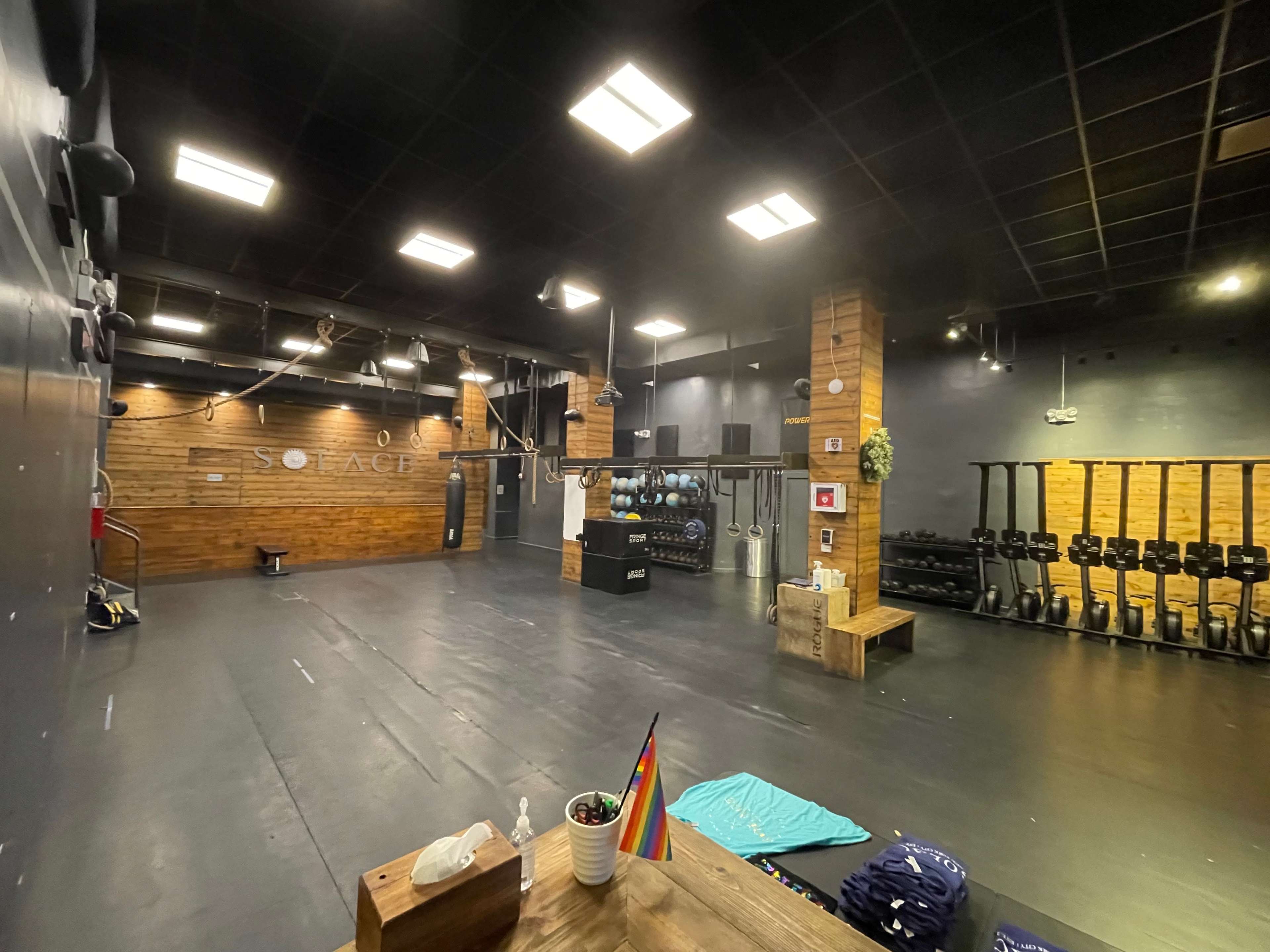 An industrial-style gym space with wooden accents, a row of rowing machines, and various fitness equipment arranged on a black floor under bright lights.