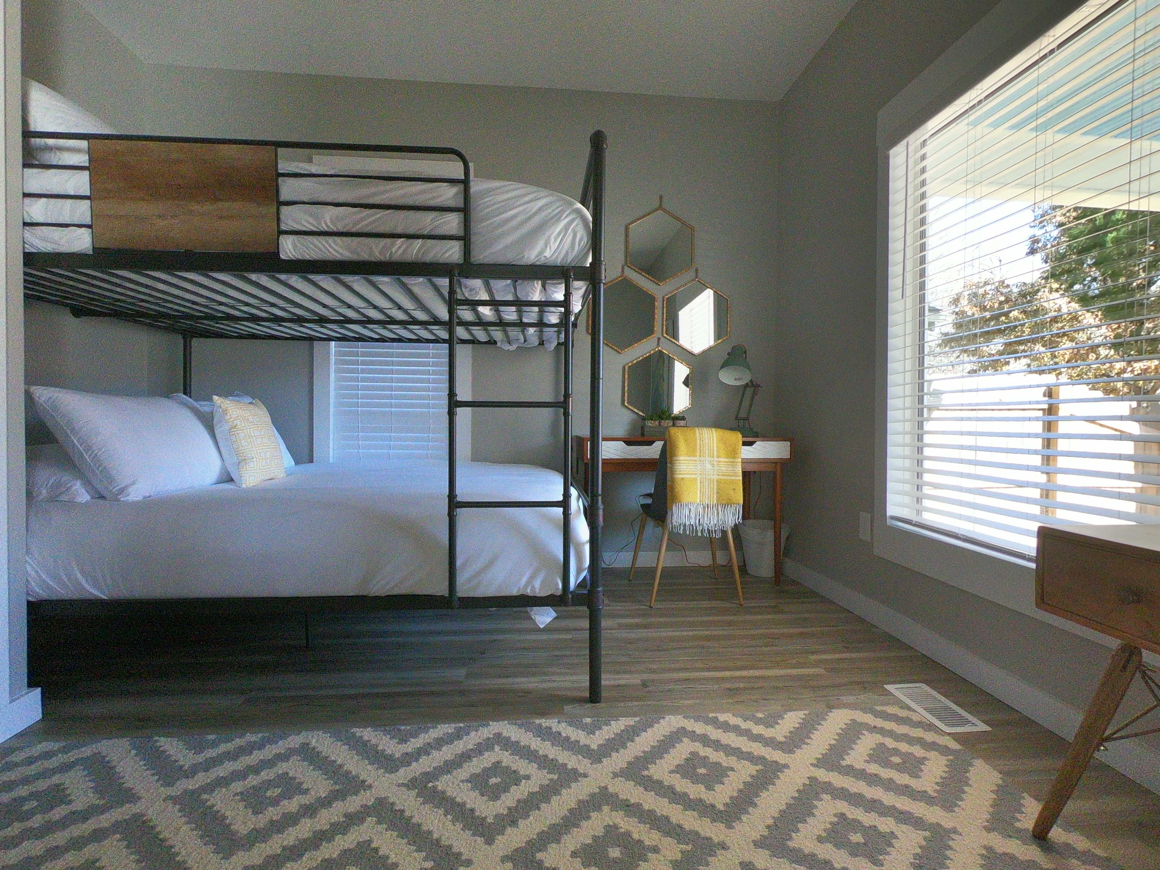 A bedroom with a bunk bed, a desk with a chair, and large windows allowing natural light to enter.