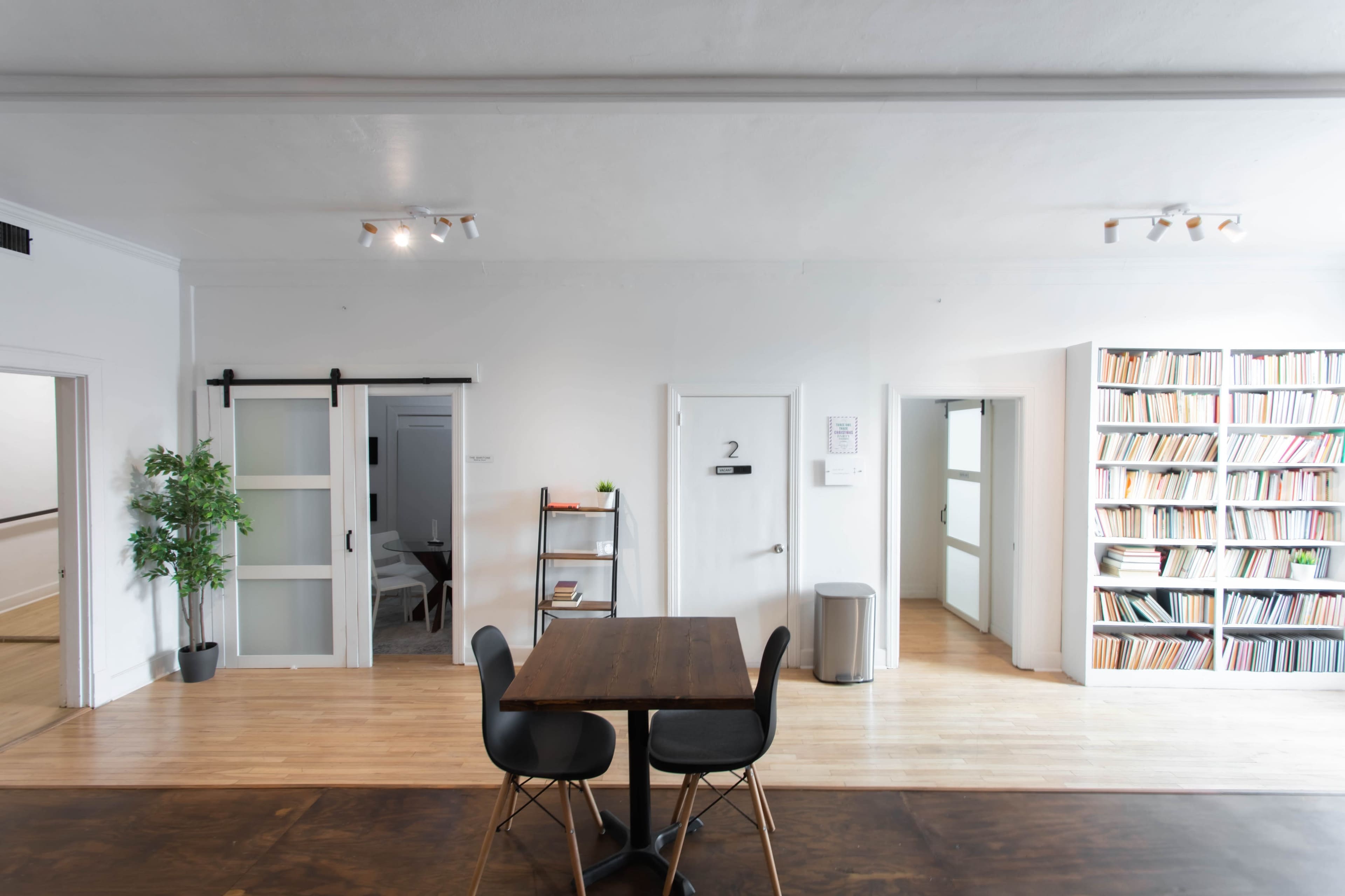 A bright room features a wooden table with black chairs, a bookshelf filled with documents, and two doorways leading to separate areas.