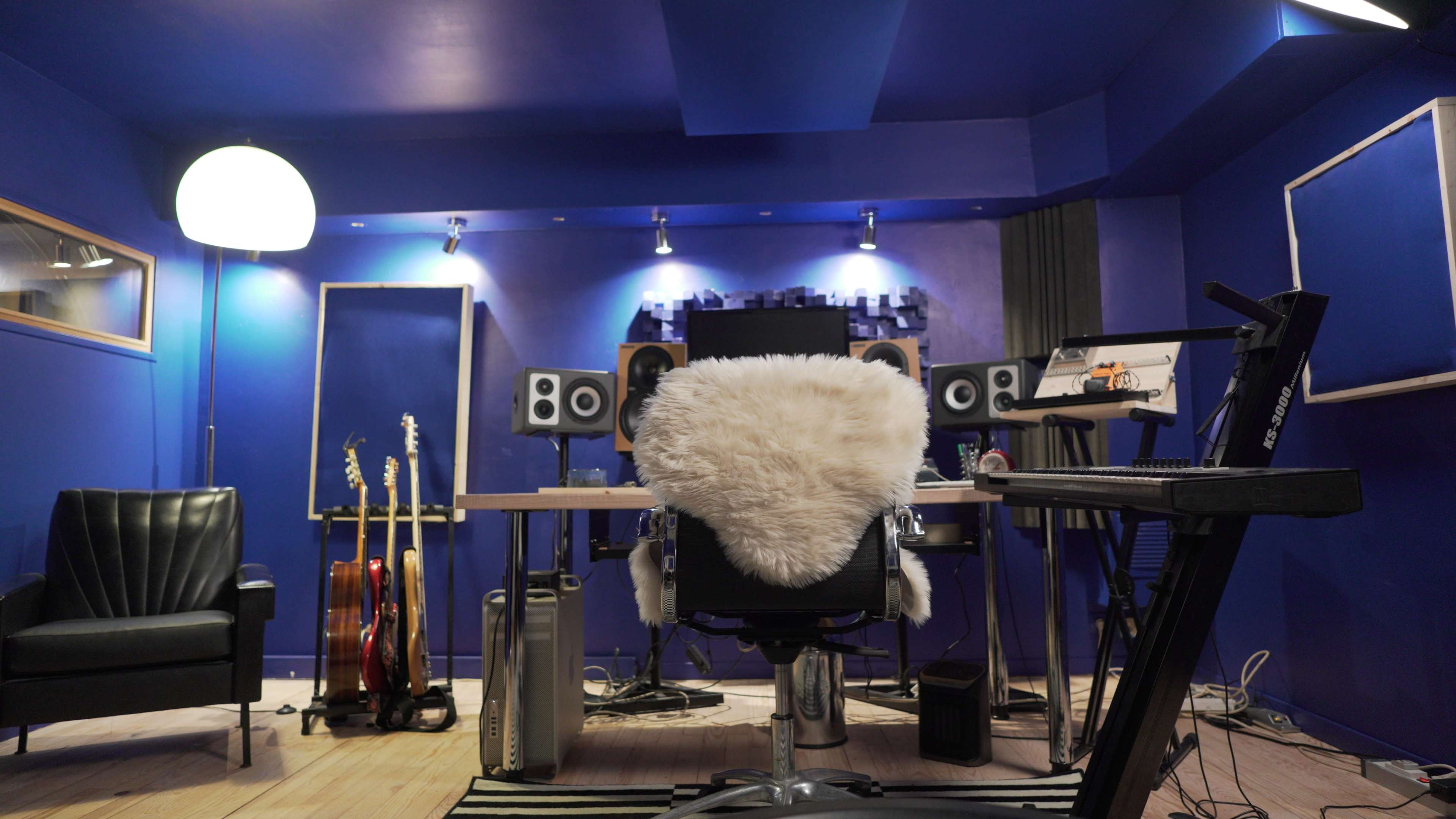 The image shows a music studio featuring a central desk with speakers, a computer monitor, and a plush chair, surrounded by instruments and a blue-painted wall.