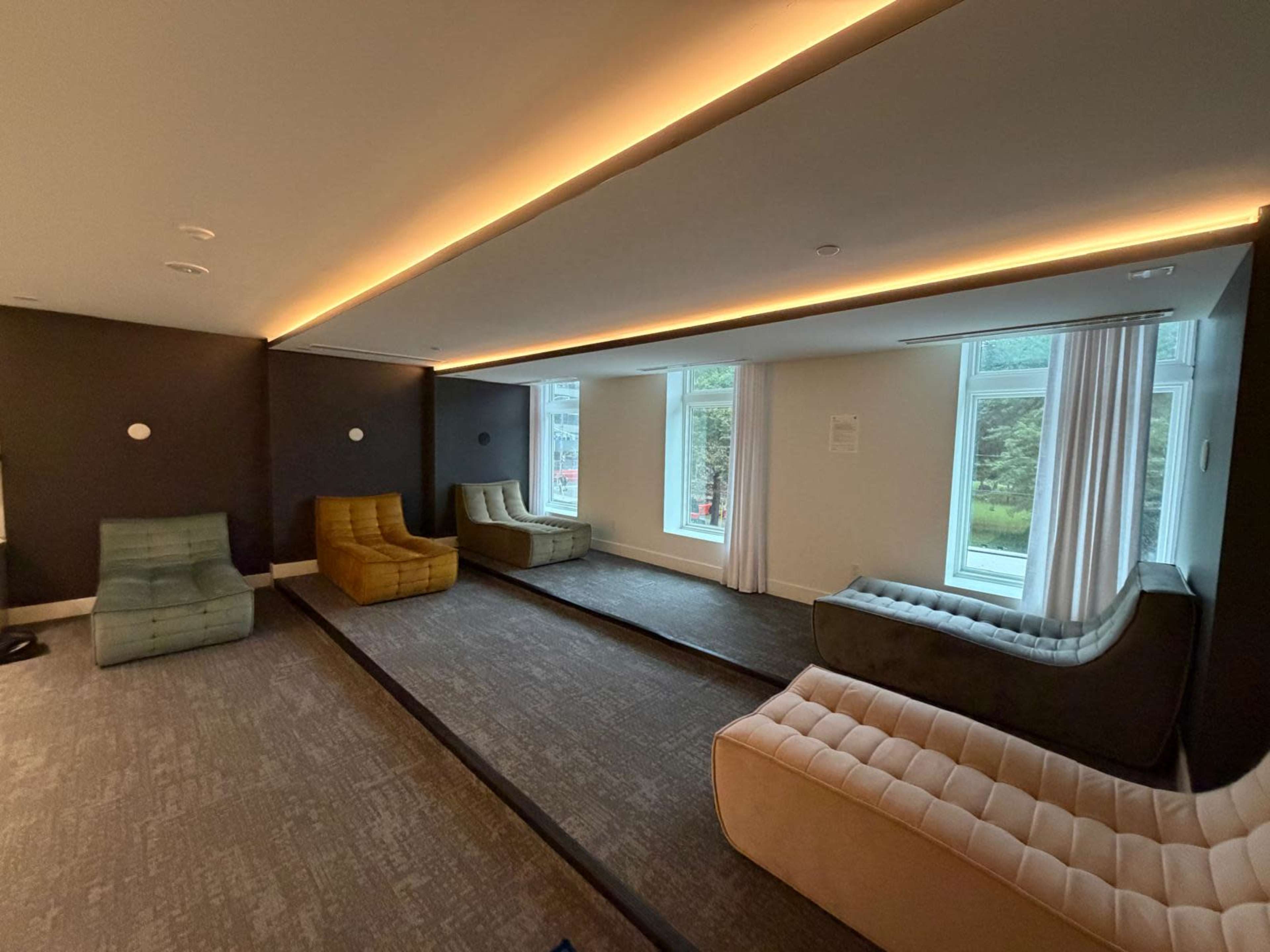 The image shows a modern lounge area with several upholstered chairs arranged on a carpeted floor, illuminated by overhead lighting and large windows letting in natural light.