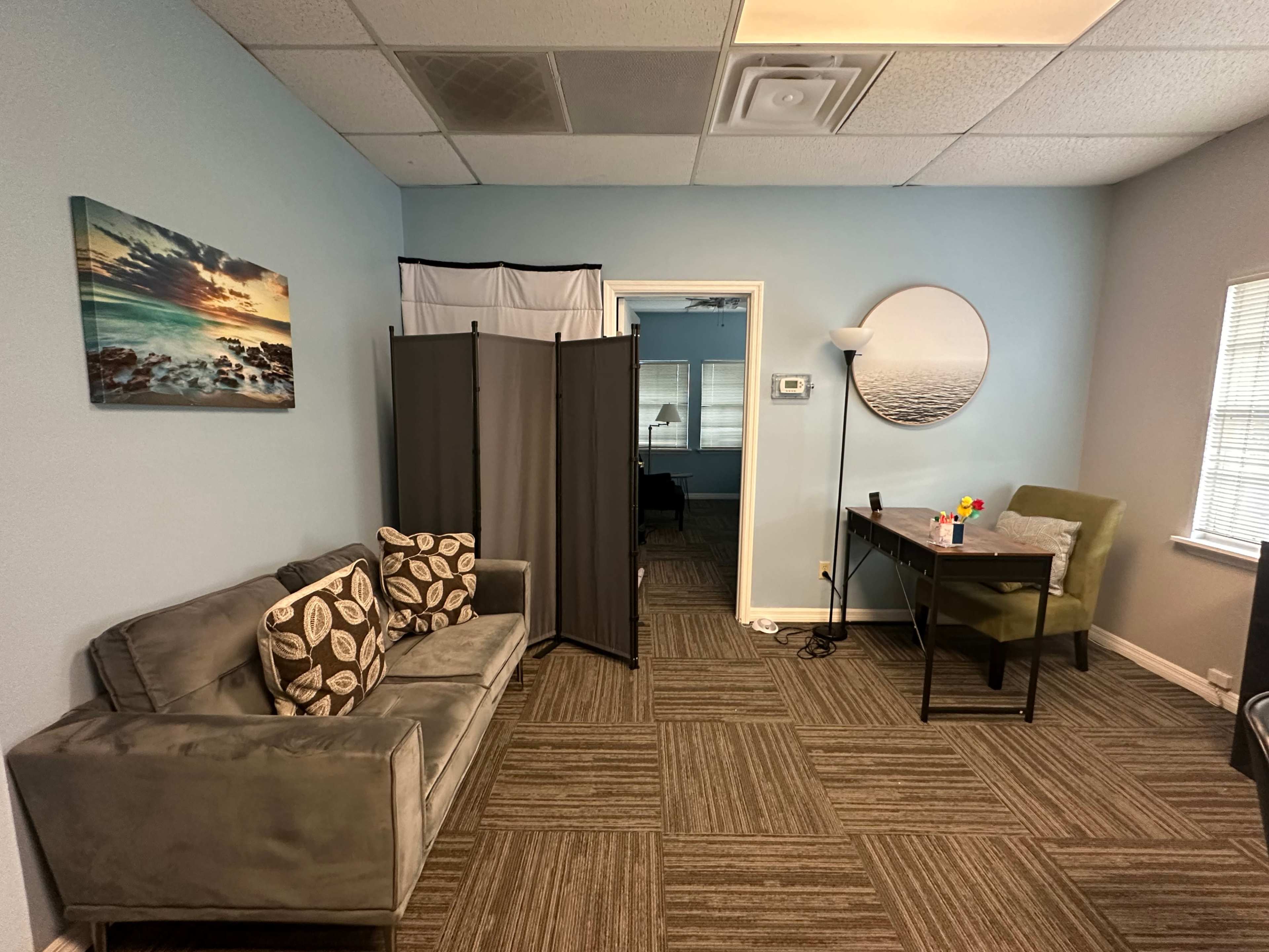 The image shows a light blue room featuring a gray couch with patterned pillows, a folding screen, a table with green chairs, and framed artwork on the walls.