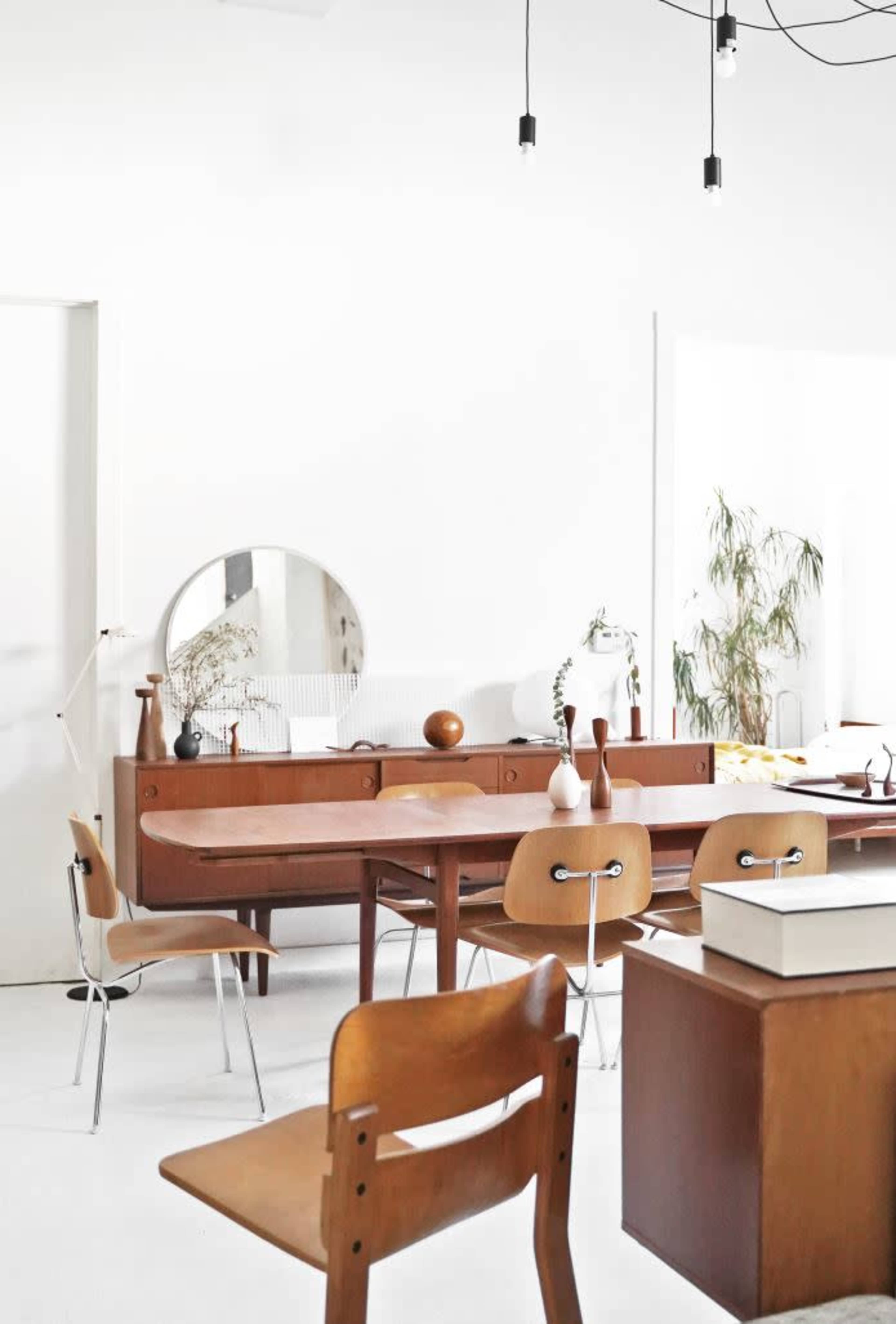 A bright, minimalist interior featuring a wooden dining table surrounded by chairs, with a large mirror and various decorative items on display.