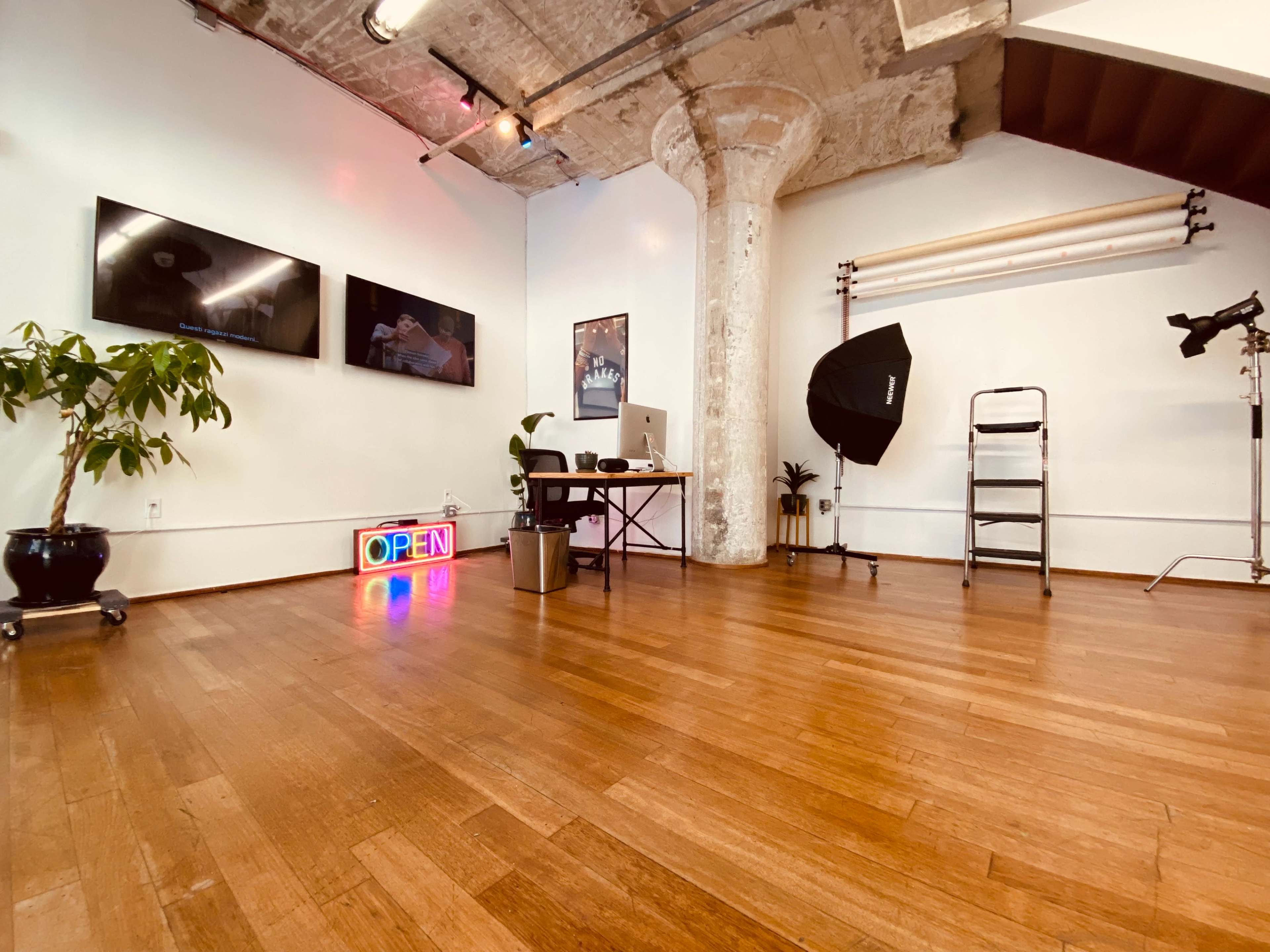 A bright, spacious studio workspace with hardwood floors, framed screens on the wall, and a neon "OPEN" sign, along with a plant and various photography equipment.