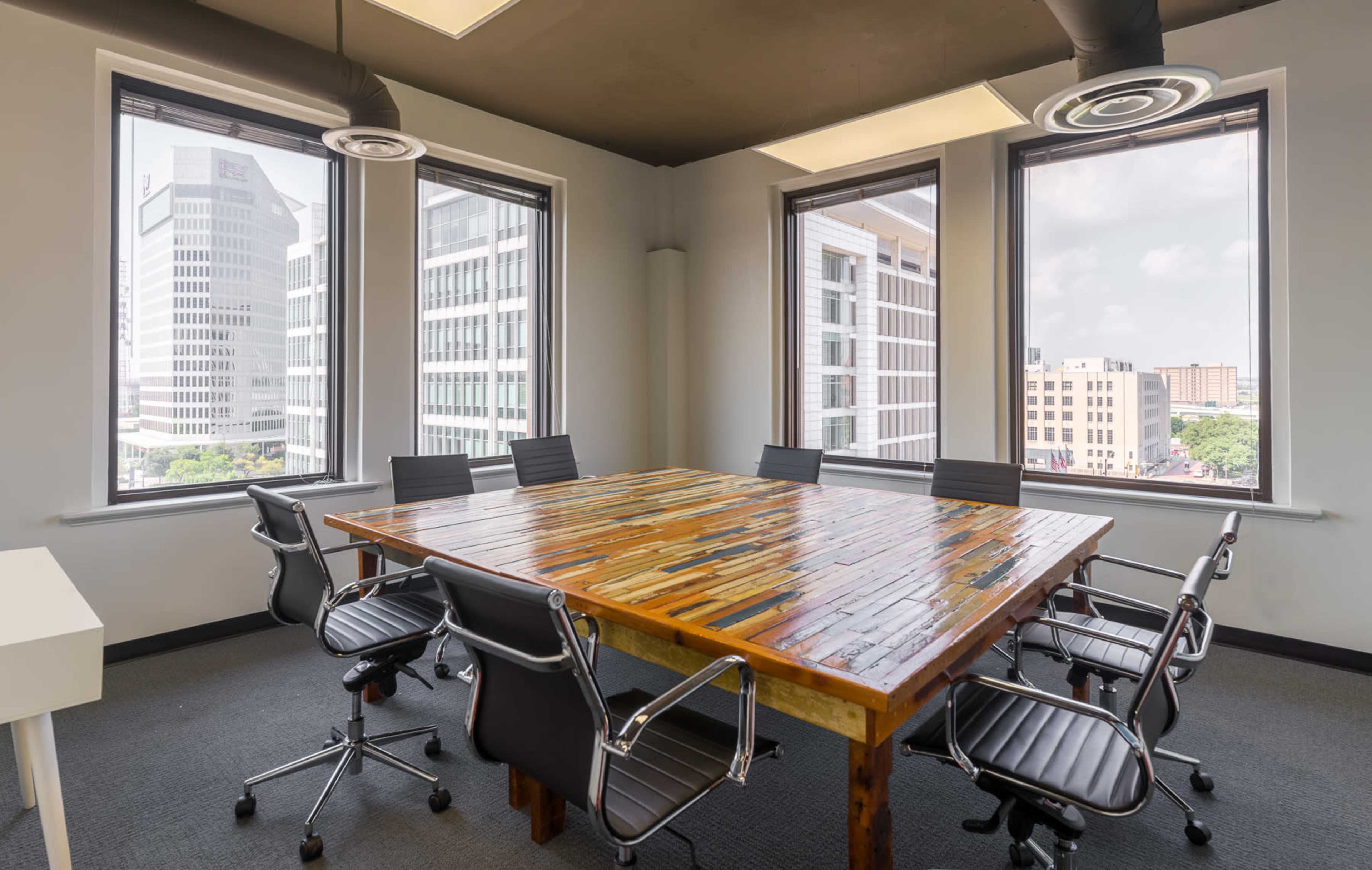 A conference room features a large, multi-colored wooden table surrounded by black office chairs with a backdrop of city skyline visible through multiple windows.