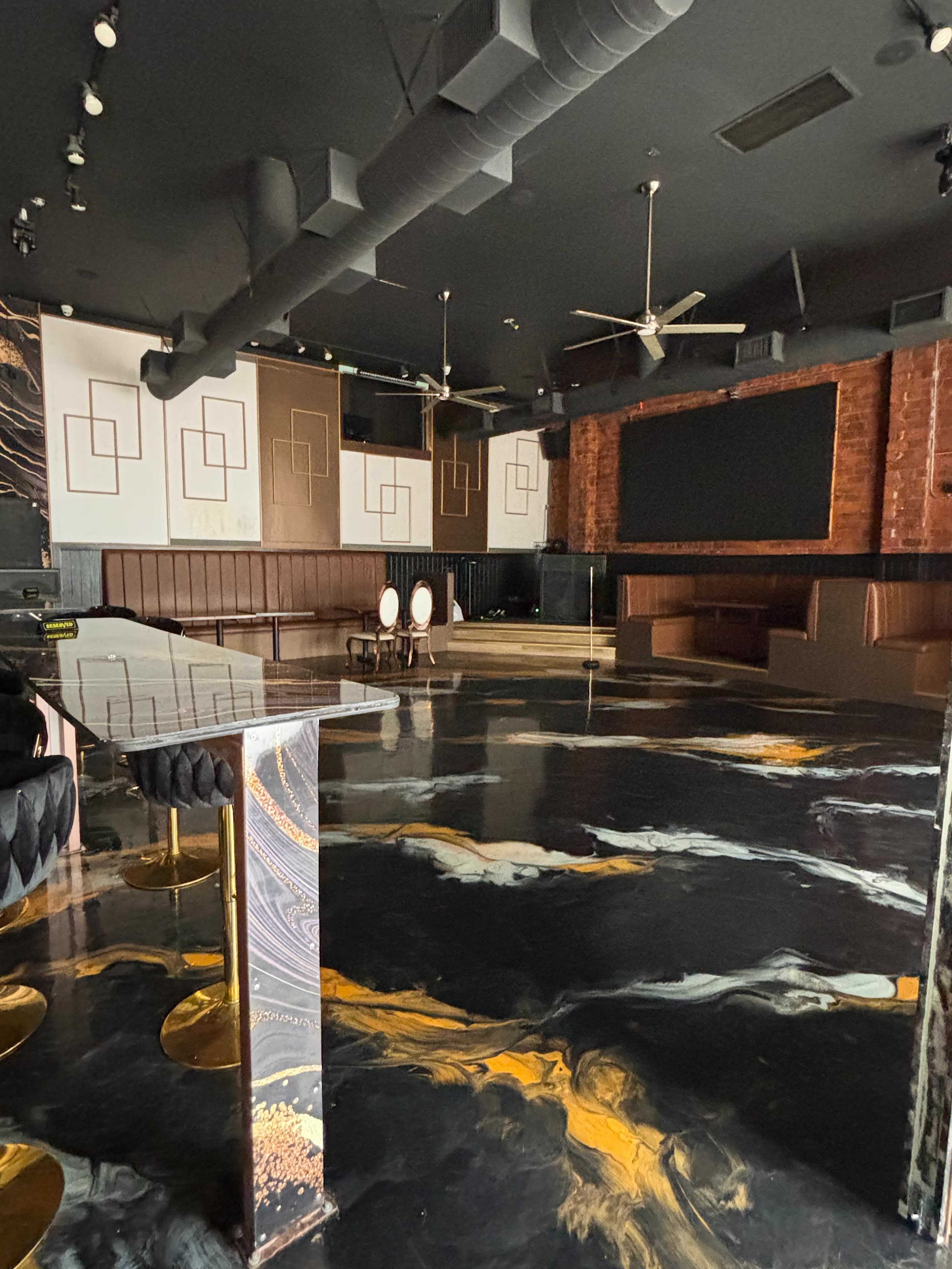 The image shows a modern lounge space with a glossy black floor featuring orange and gold patterns, complementing a stylish seating area and a large screen on one wall.
