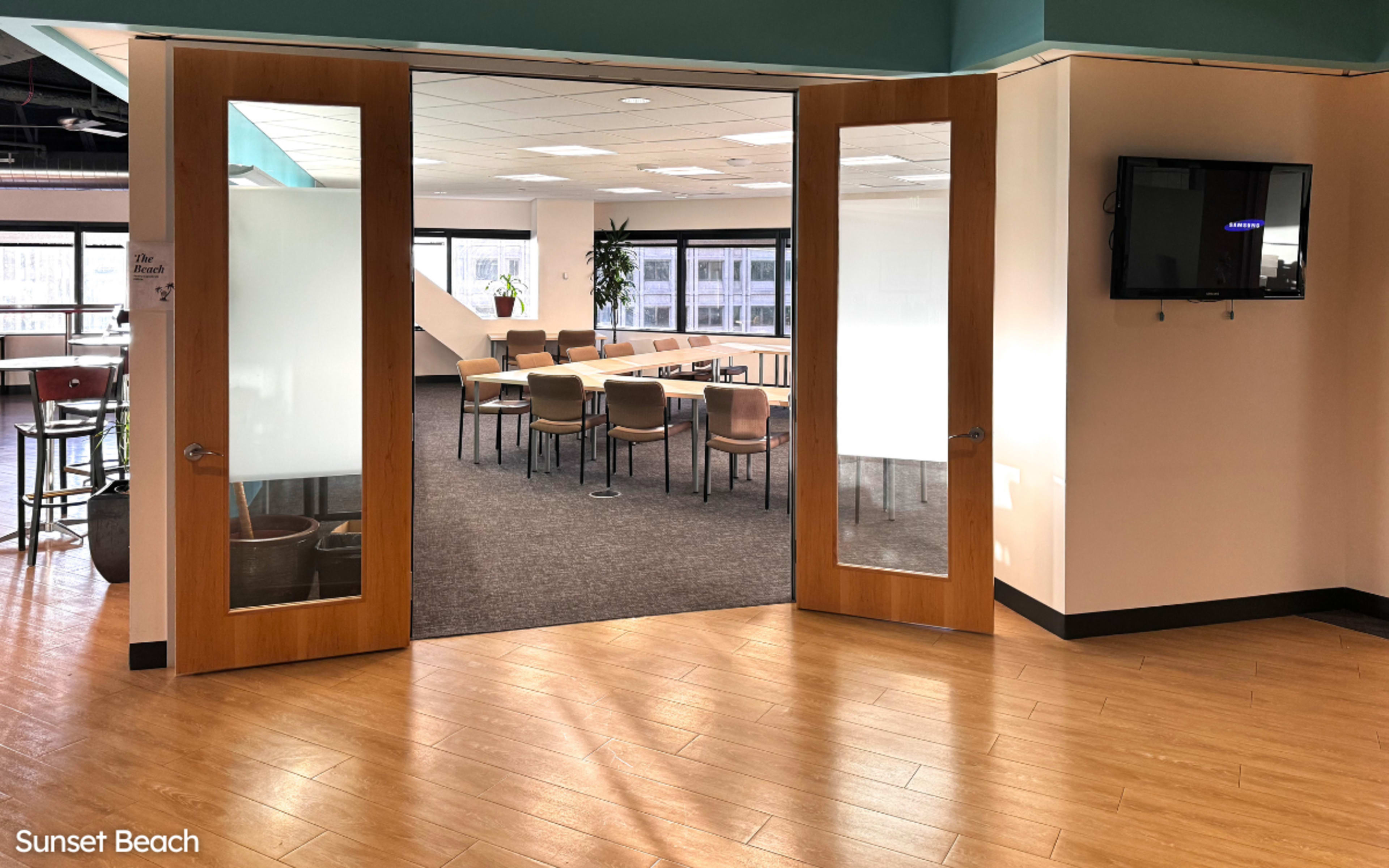 A meeting room with a large table surrounded by chairs, visible through glass double doors.