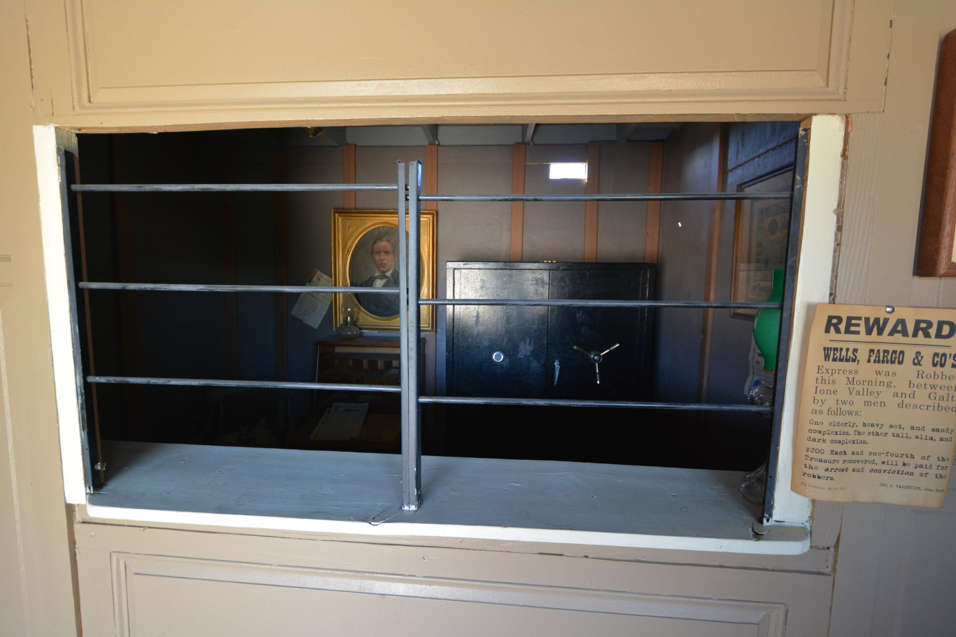 The image shows a barred window opening with a framed portrait visible in the background and a safe against the wall.
