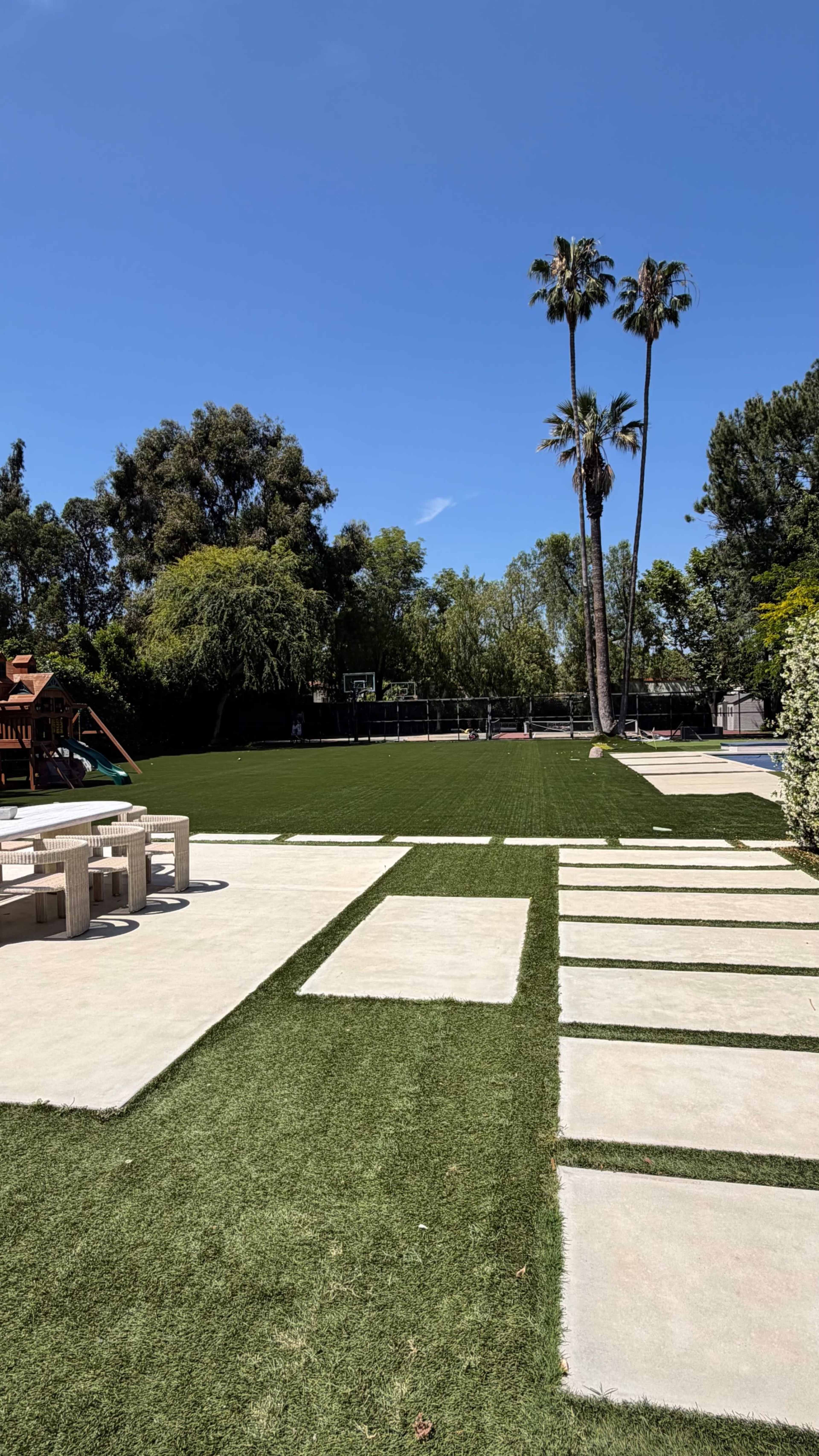 A spacious backyard features artificial grass, concrete pathways, palm trees, and an area with a playground set.