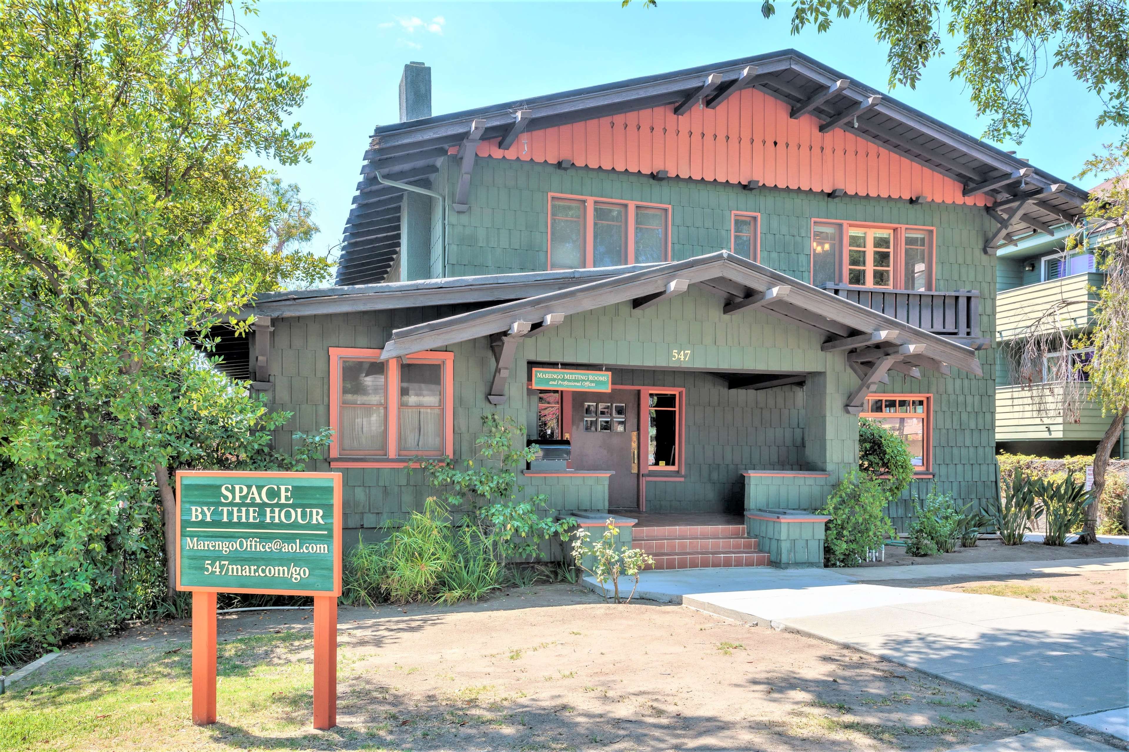 A large green wooden house with a red-trimmed gabled roof, featuring an inviting entrance and a sign indicating it is available for rent by the hour.