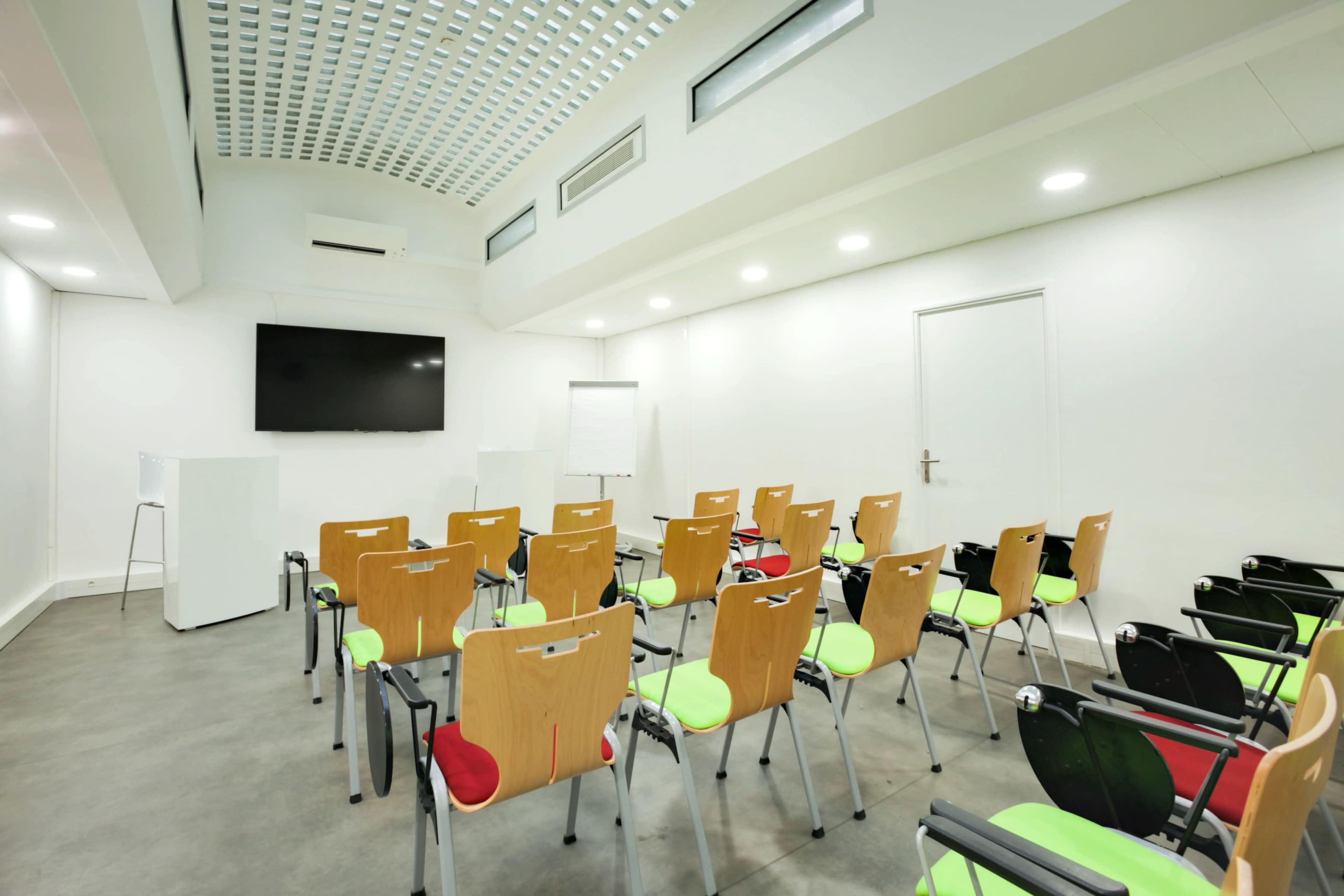 A modern meeting room with rows of wooden chairs featuring green and red seats, a large screen at the front, and a whiteboard.