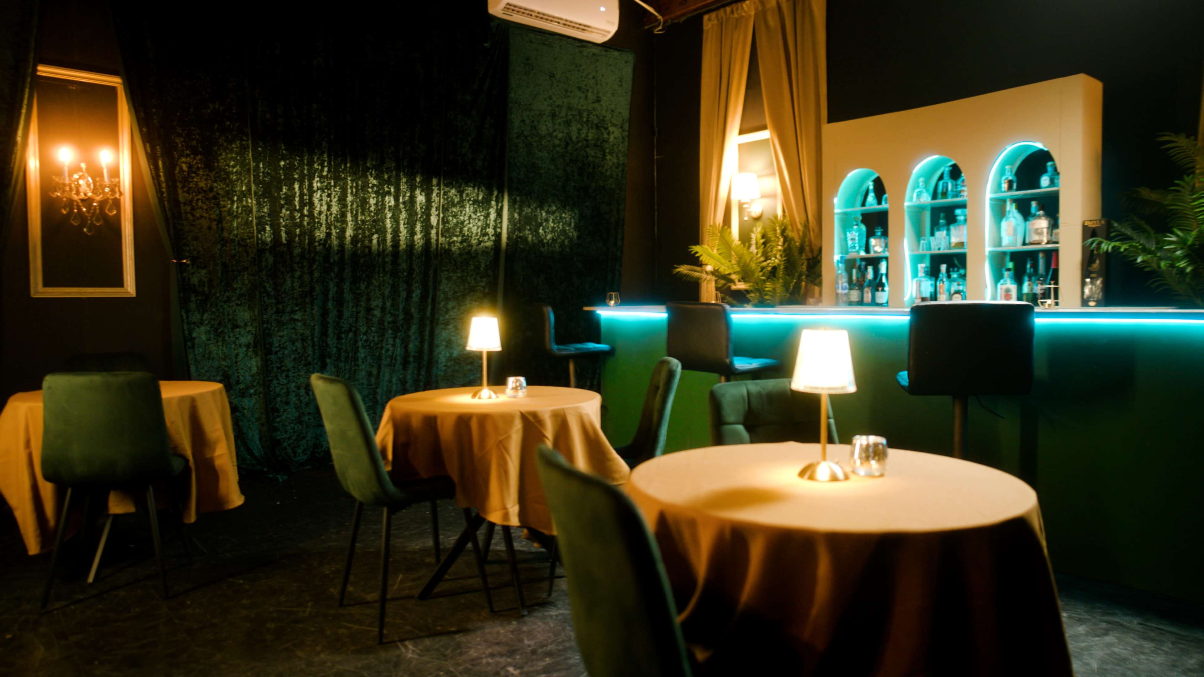 The image shows a dimly lit bar area with tables covered in beige cloths, green chairs, and a well-lit back bar displaying bottles.