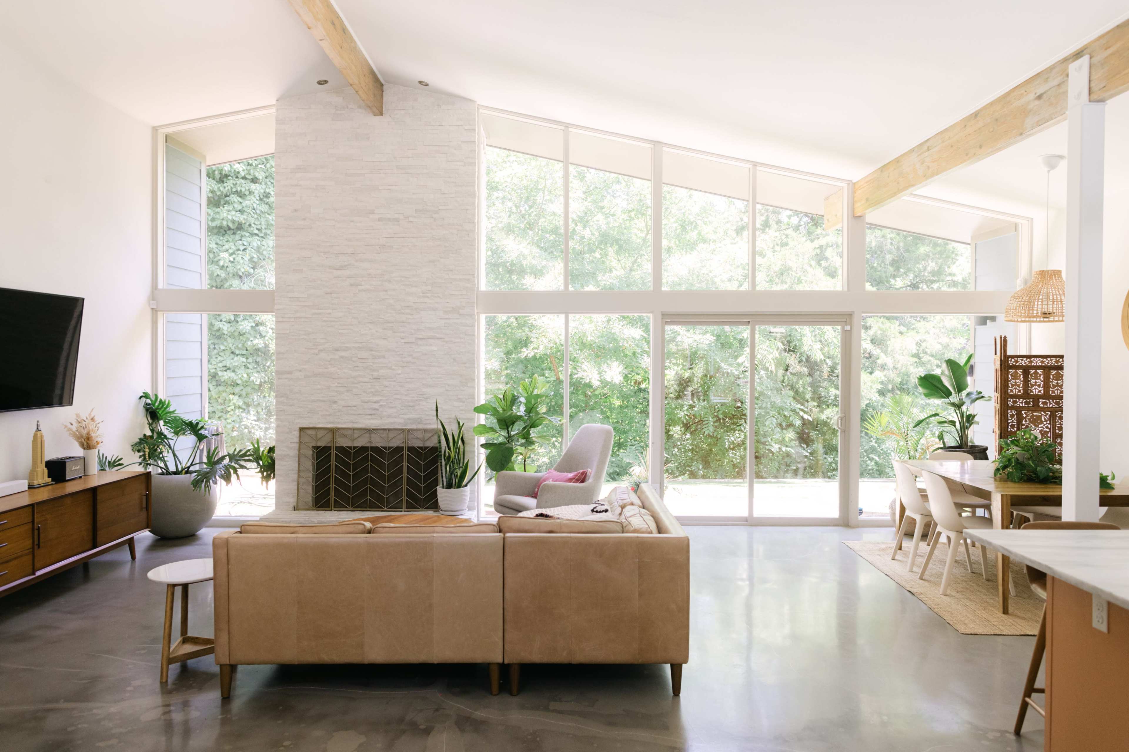 A bright living room with large windows overlooking a green landscape, featuring a beige sofa, a stone fireplace, and a dining area.