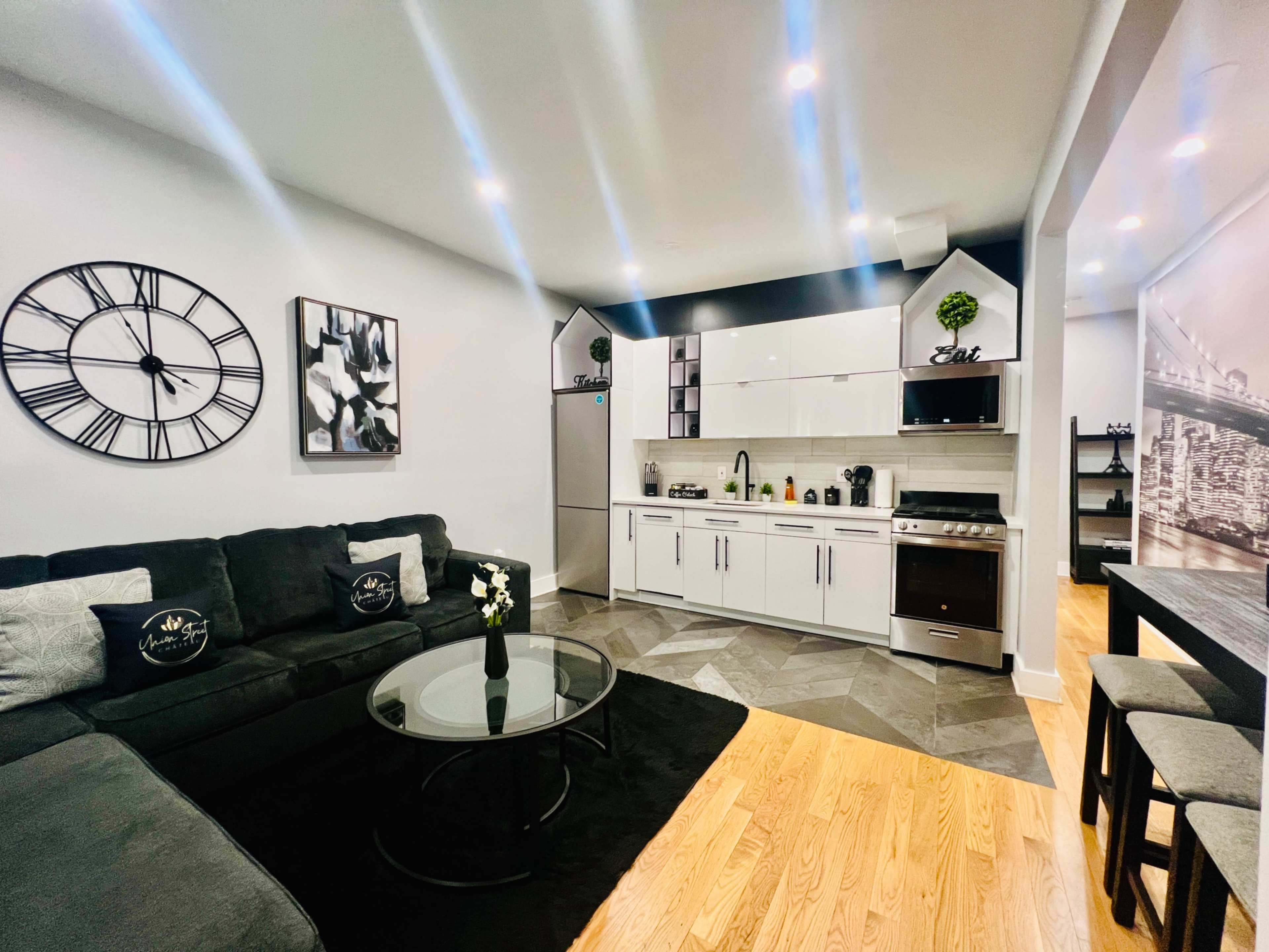 The image shows a modern living area featuring a sectional couch, a glass coffee table, a kitchen with sleek white cabinetry, and decorative wall art.