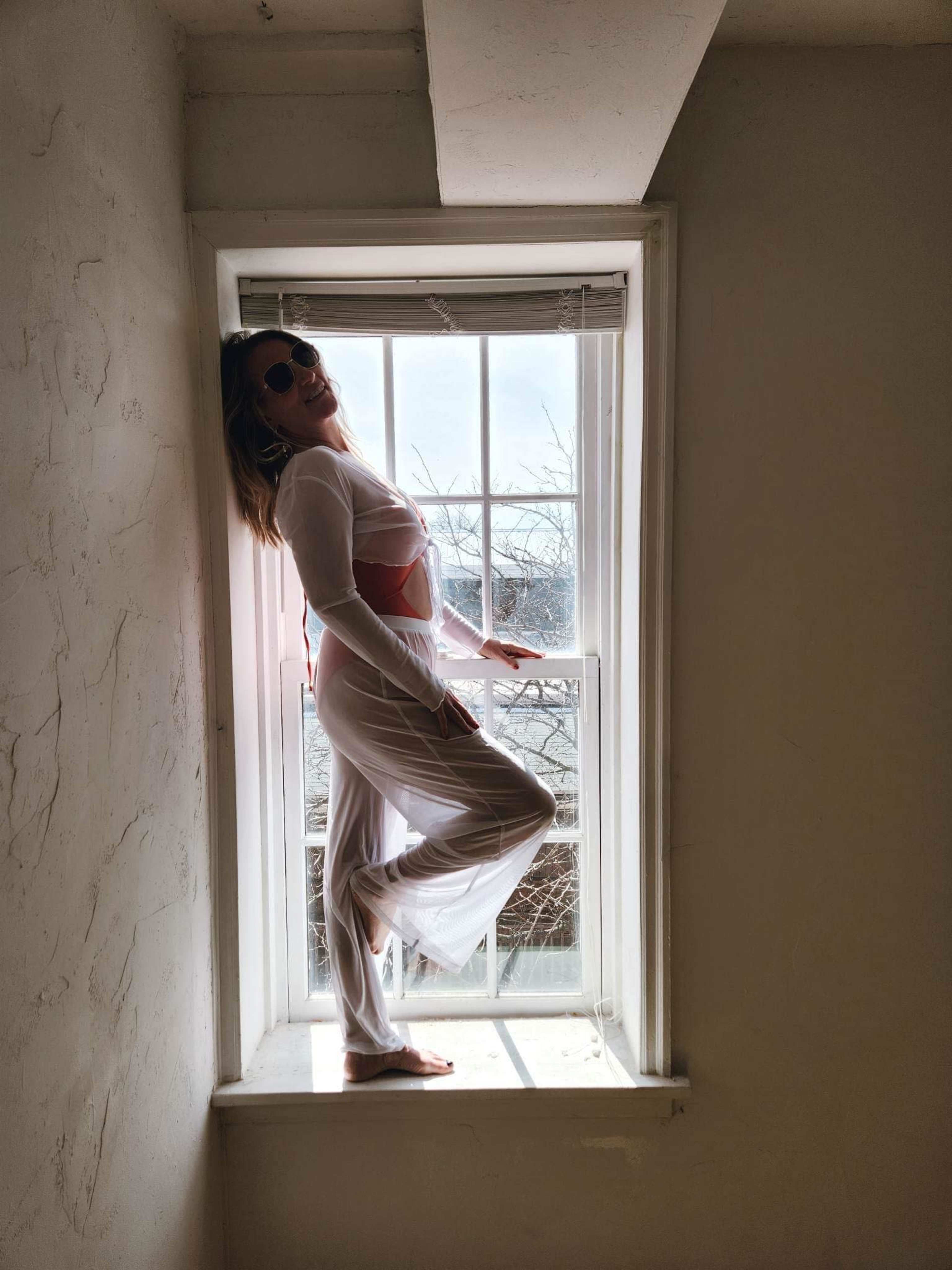 A person stands in a window nook, striking a pose while wearing a sheer outfit and sunglasses.