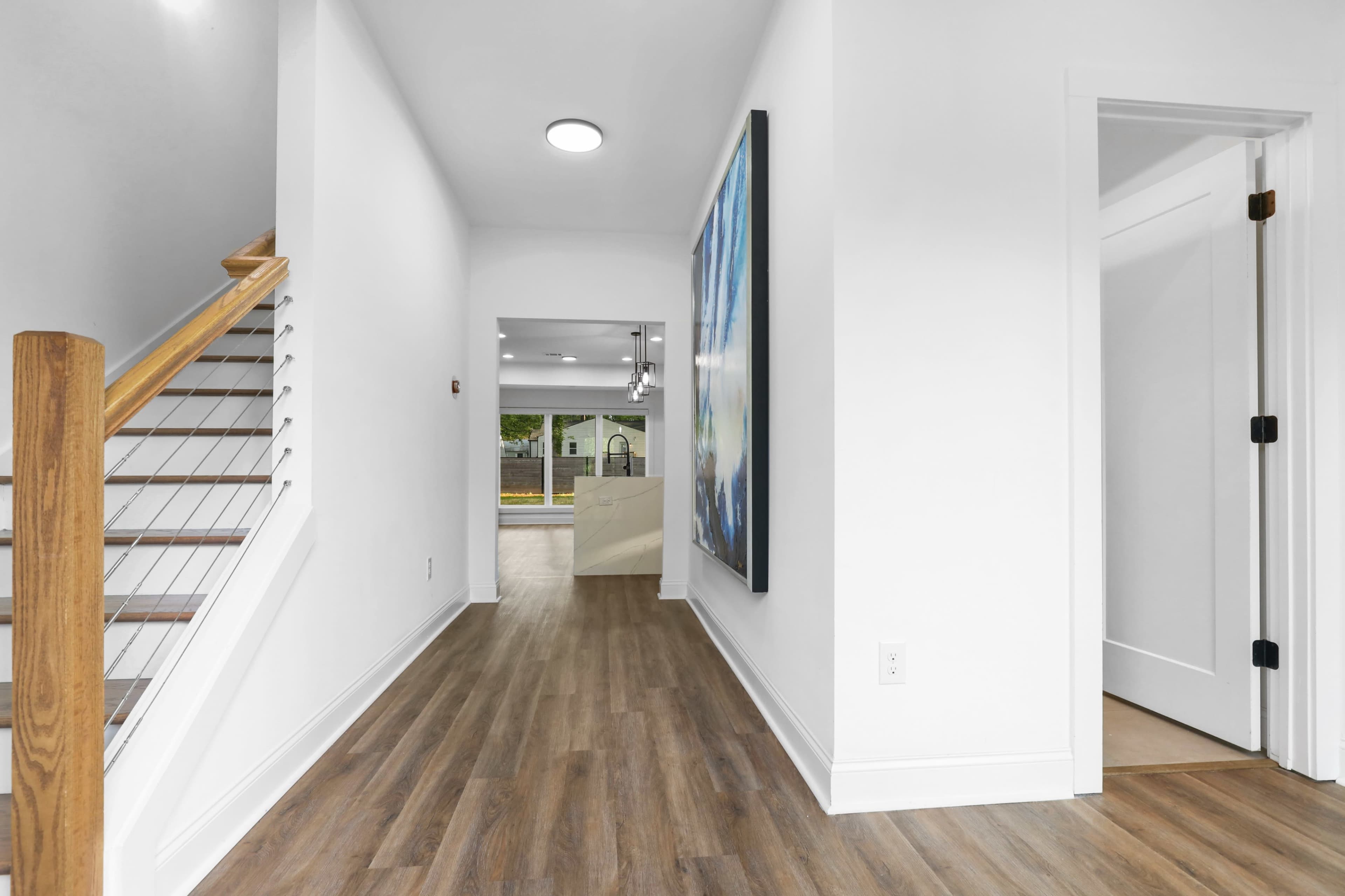 A modern hallway features a wooden staircase on the left, with a large artwork hanging on the right wall and doors leading to other rooms at the end.