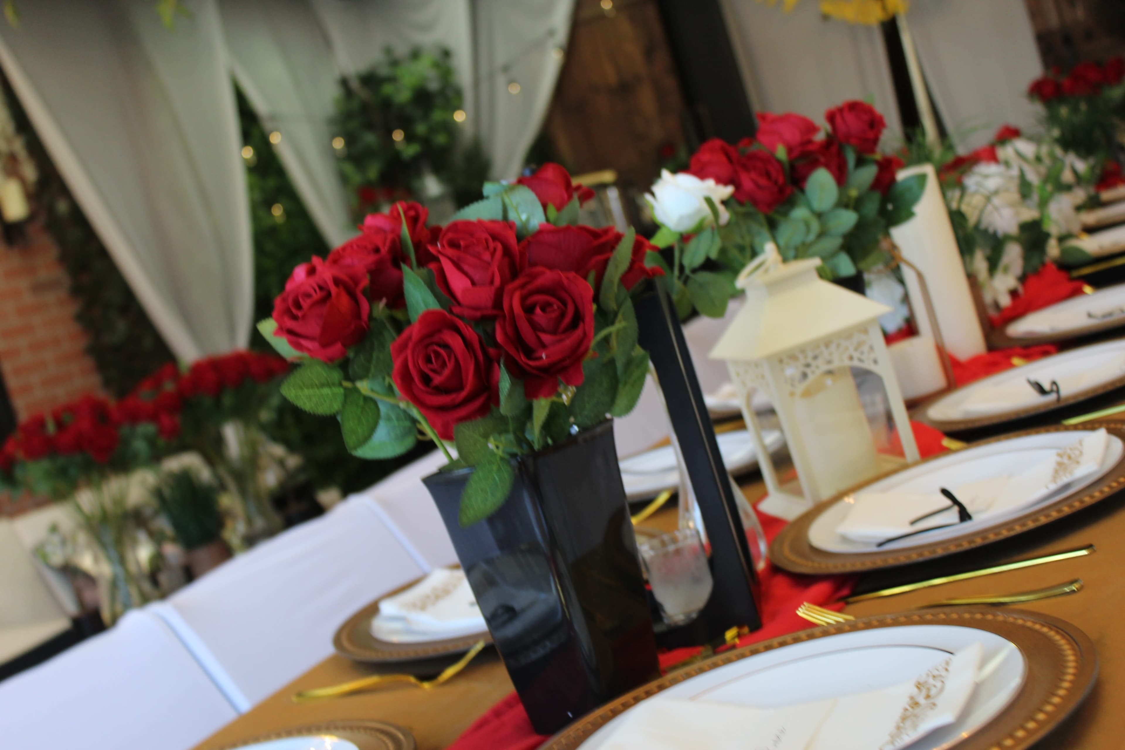 A table is elegantly set with plates, cutlery, and decorative vases of red roses, surrounded by soft lighting and draped fabric.