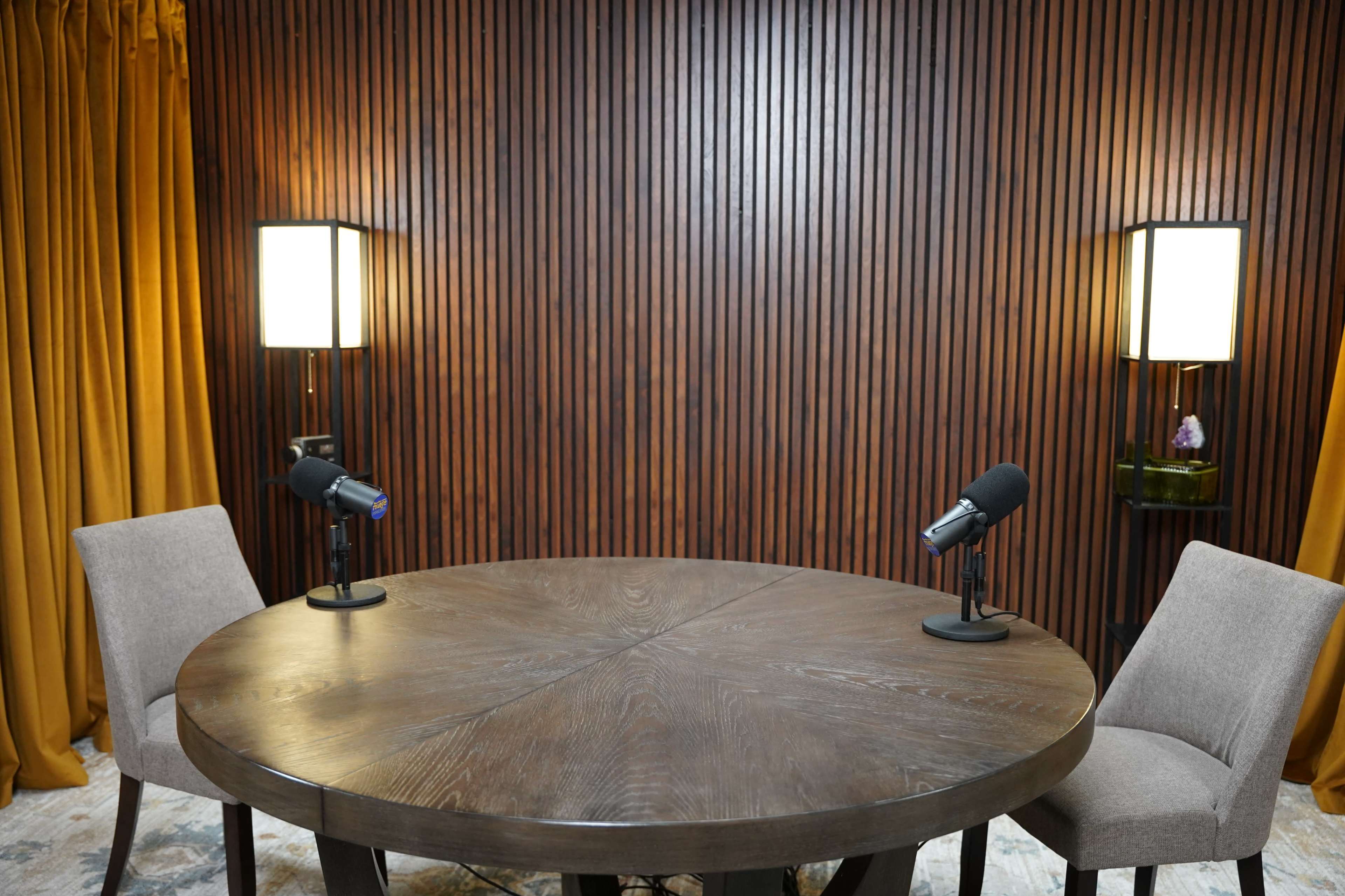 A round wooden table with two microphones is set between two chairs in front of a paneled wall and flanked by lit lamps.