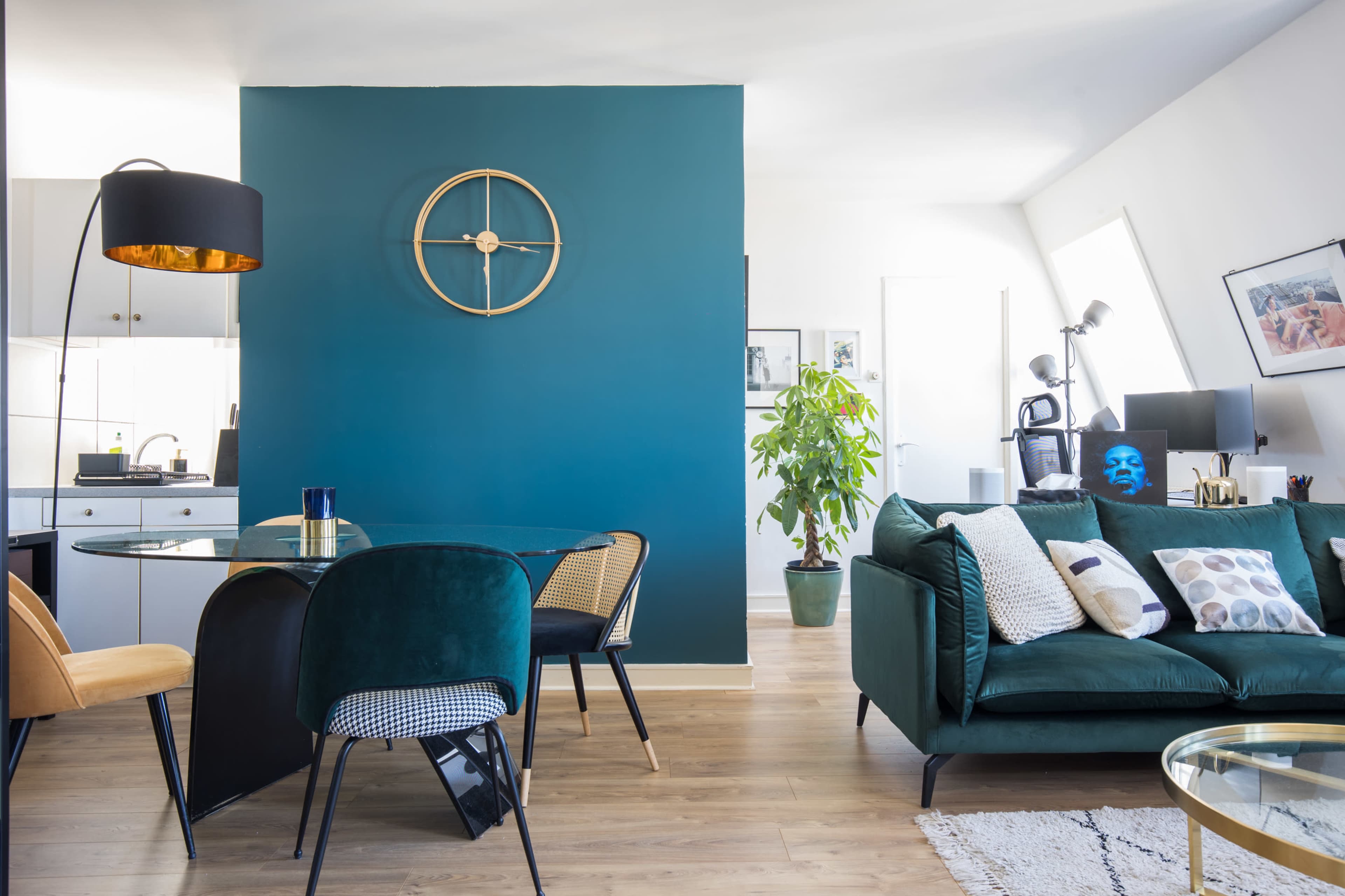 A modern living space features a green accent wall, a circular clock, a dining table with mixed seating, and a potted plant, alongside a cozy sofa adorned with decorative pillows.