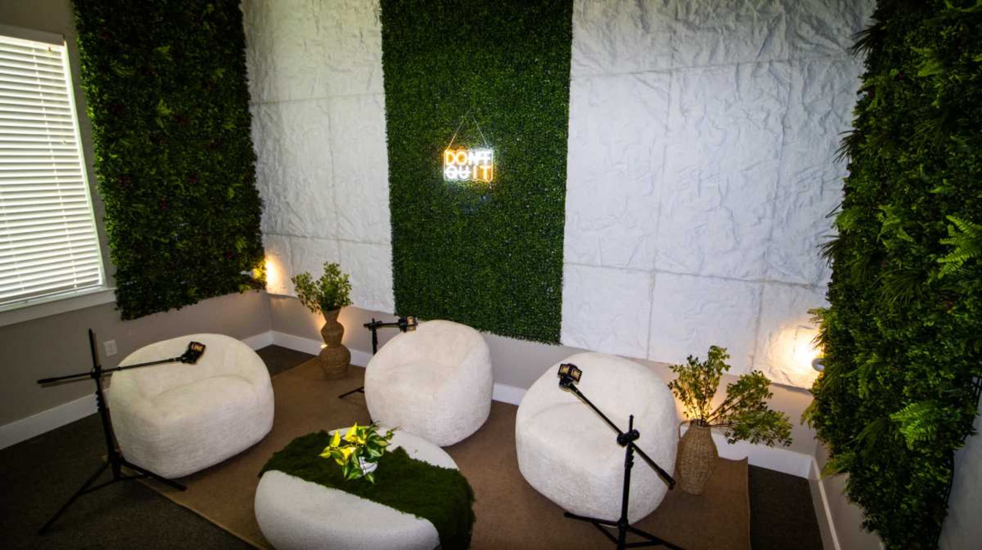 The image shows a cozy recording space featuring three plush chairs, two microphones on stands, and a circular table adorned with greenery, all set against a backdrop of textured walls and plants.