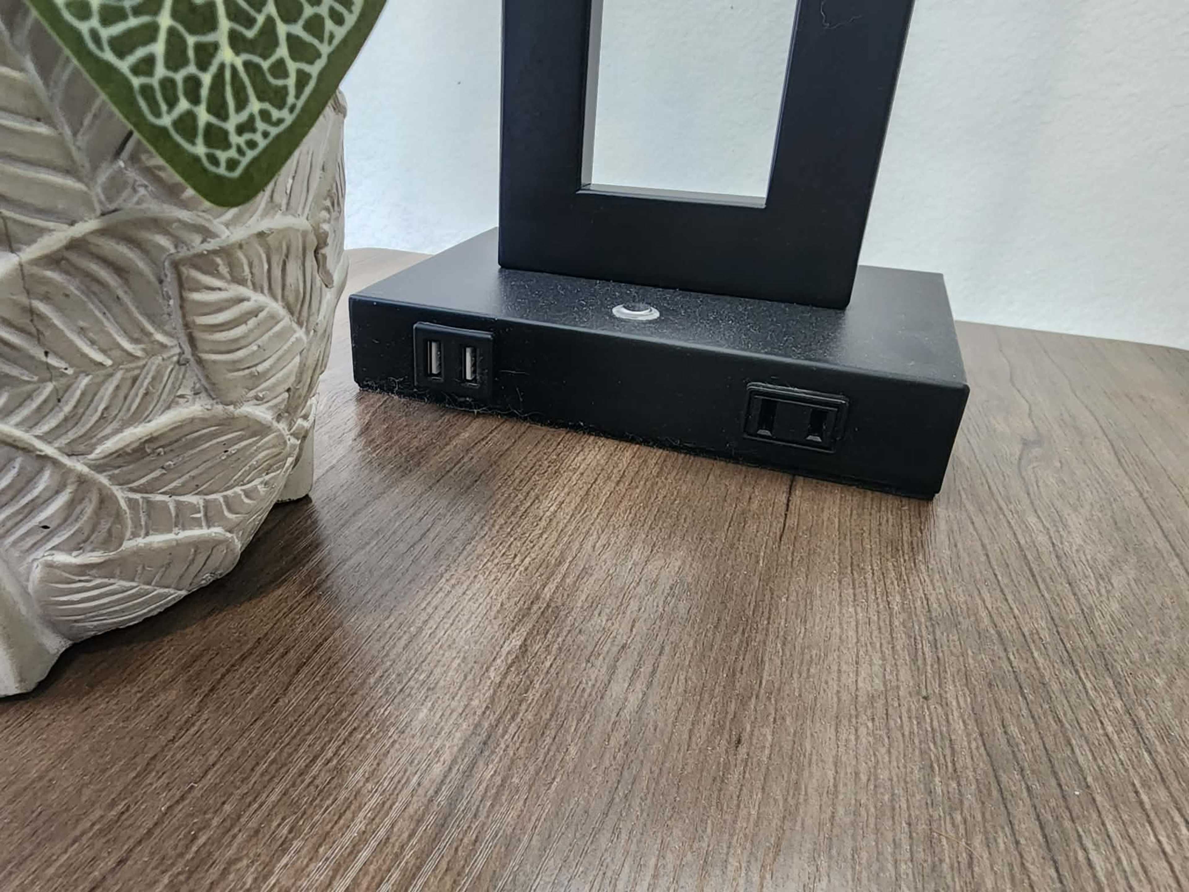 A black lamp base with two USB ports is positioned on a wooden table beside a decorative white planter.