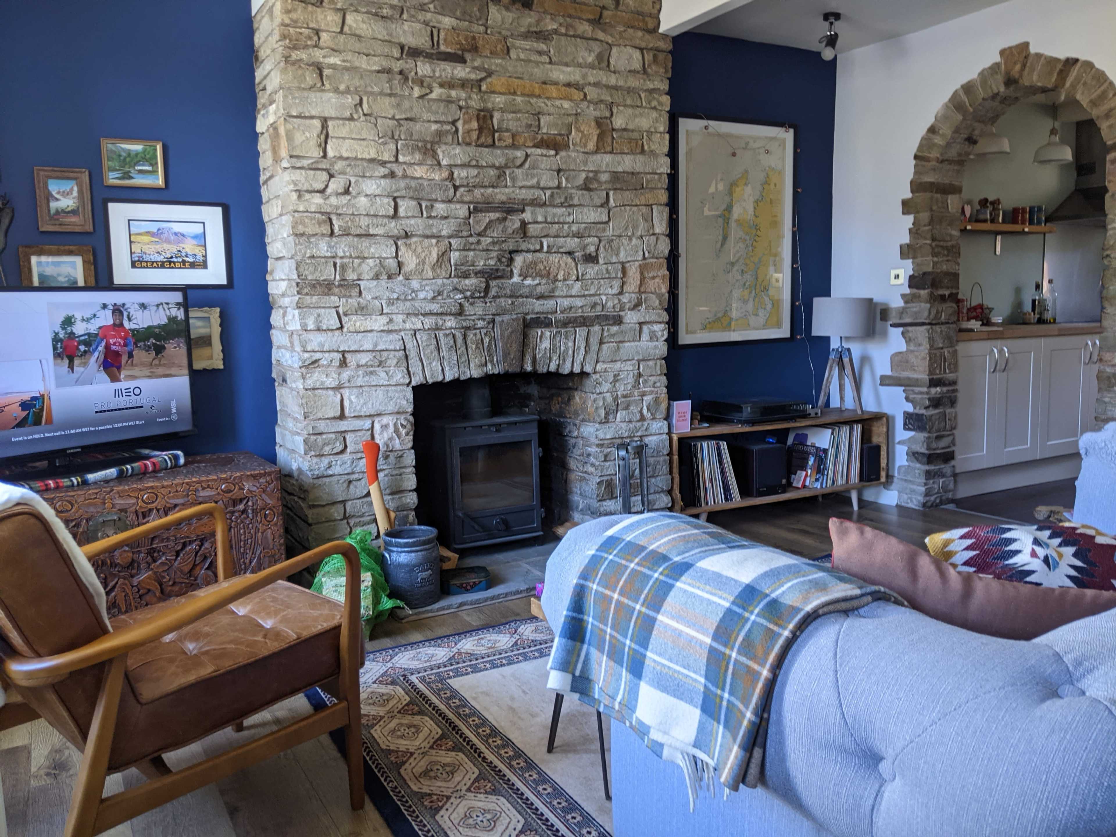A cozy living room featuring a stone fireplace, a wooden armchair, a sofa with a blanket, and shelves filled with books and decor, set against a blue wall.