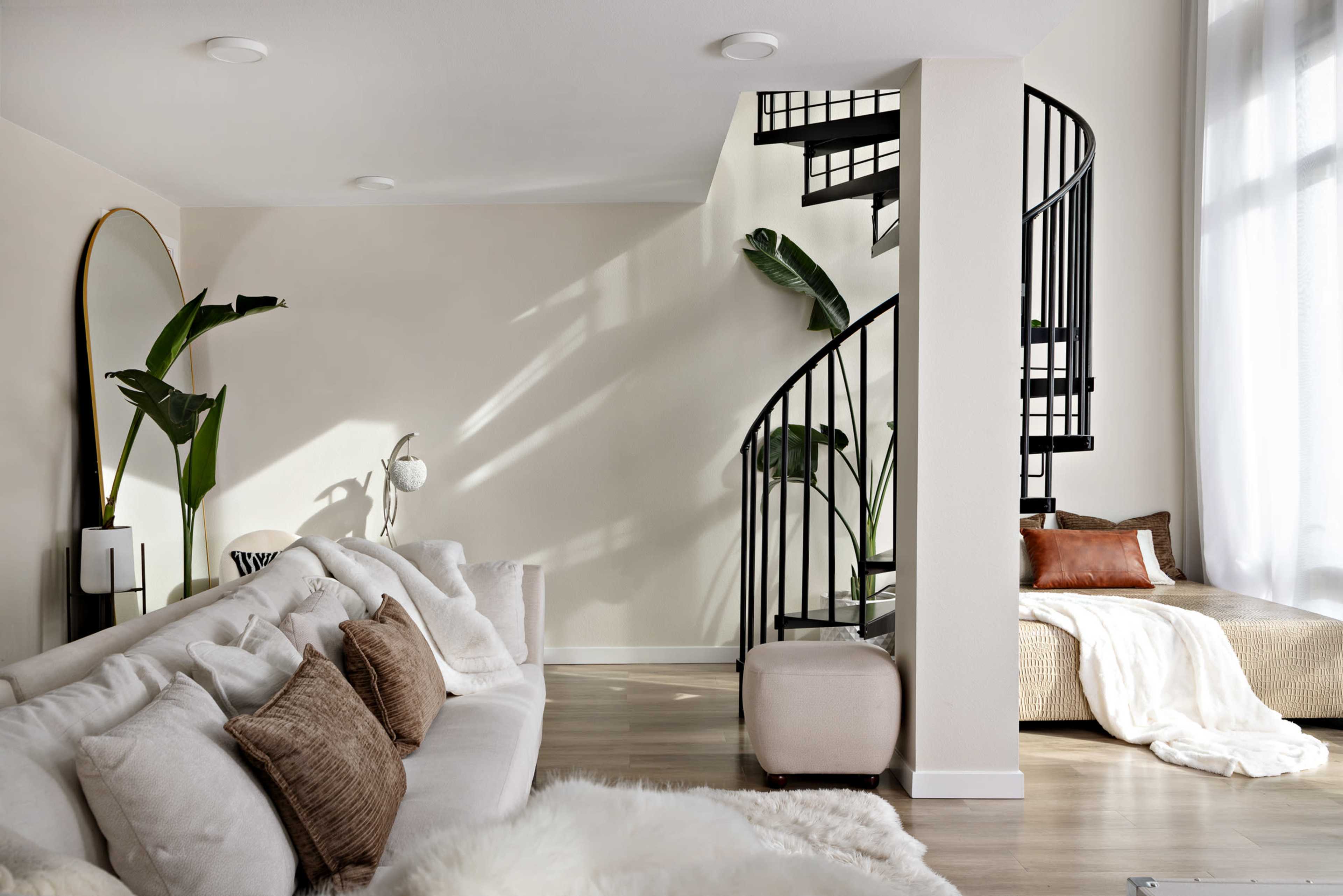 The image shows a modern living room with a spiral staircase, a large mirror, and a sofa adorned with various pillows.